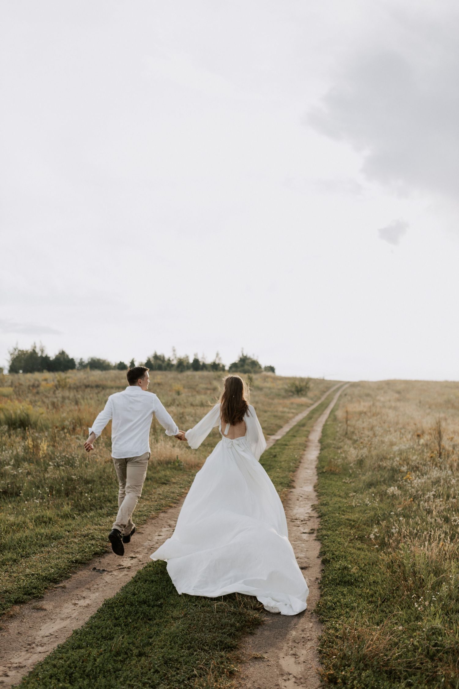 Couple in white running down dirt path through meadow during romantic photoshoot.