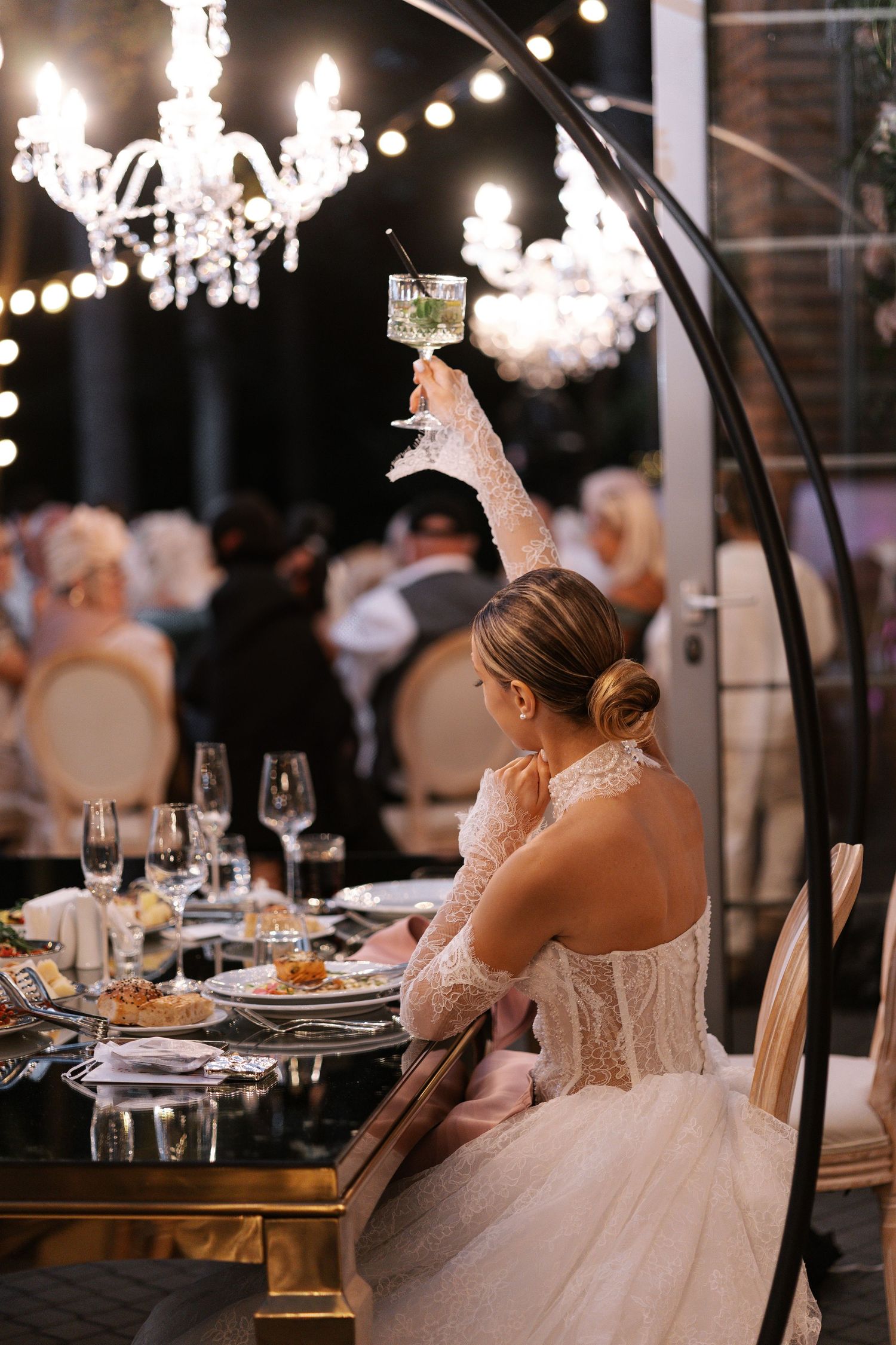 Bride in white gown raises a toast at elegantly decorated reception table under crystal chandelier.