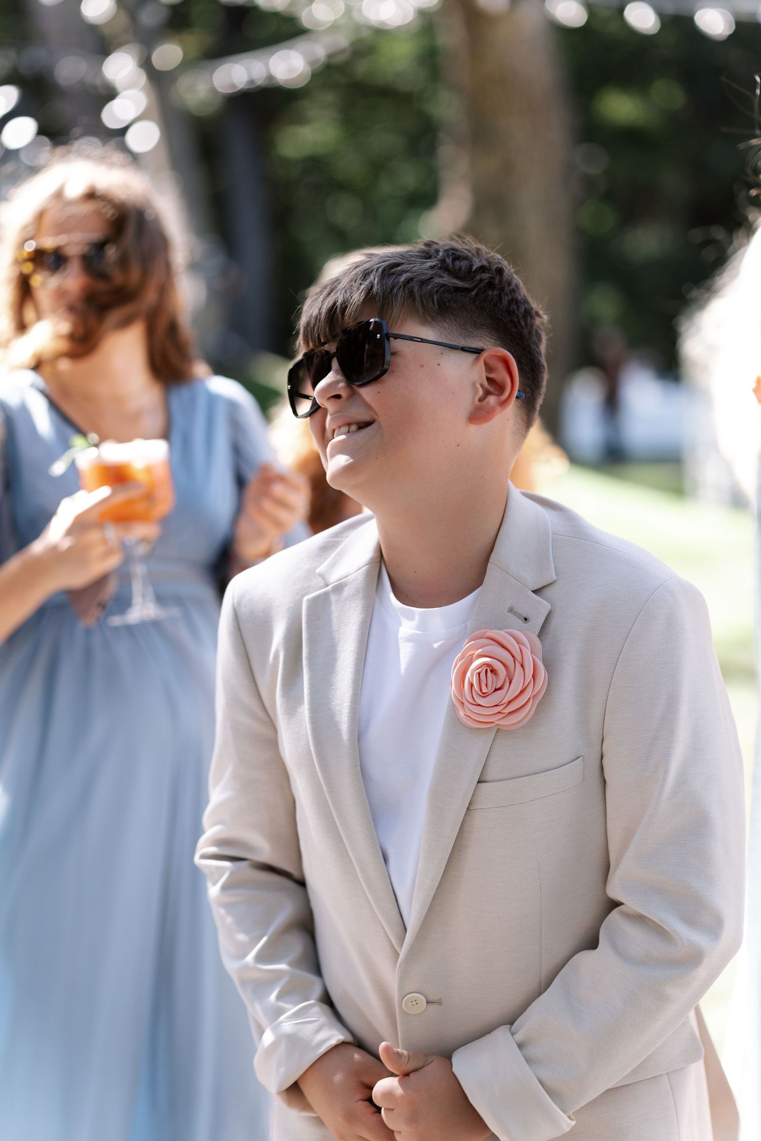Wedding guest in a light beige suit with pink rose boutonniere smiles outdoors.