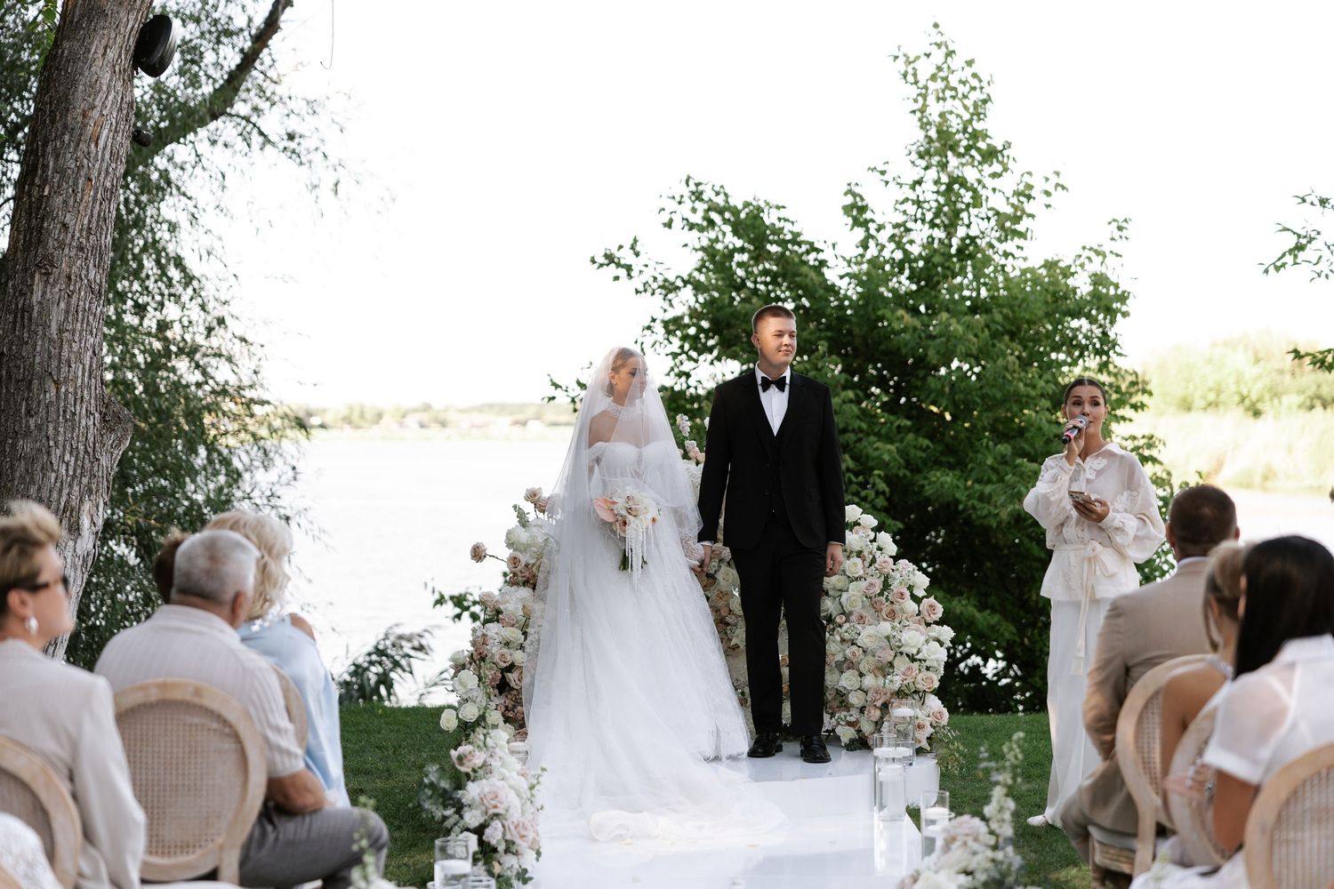 A wedding party poses outdoors in front of trees and greenery.