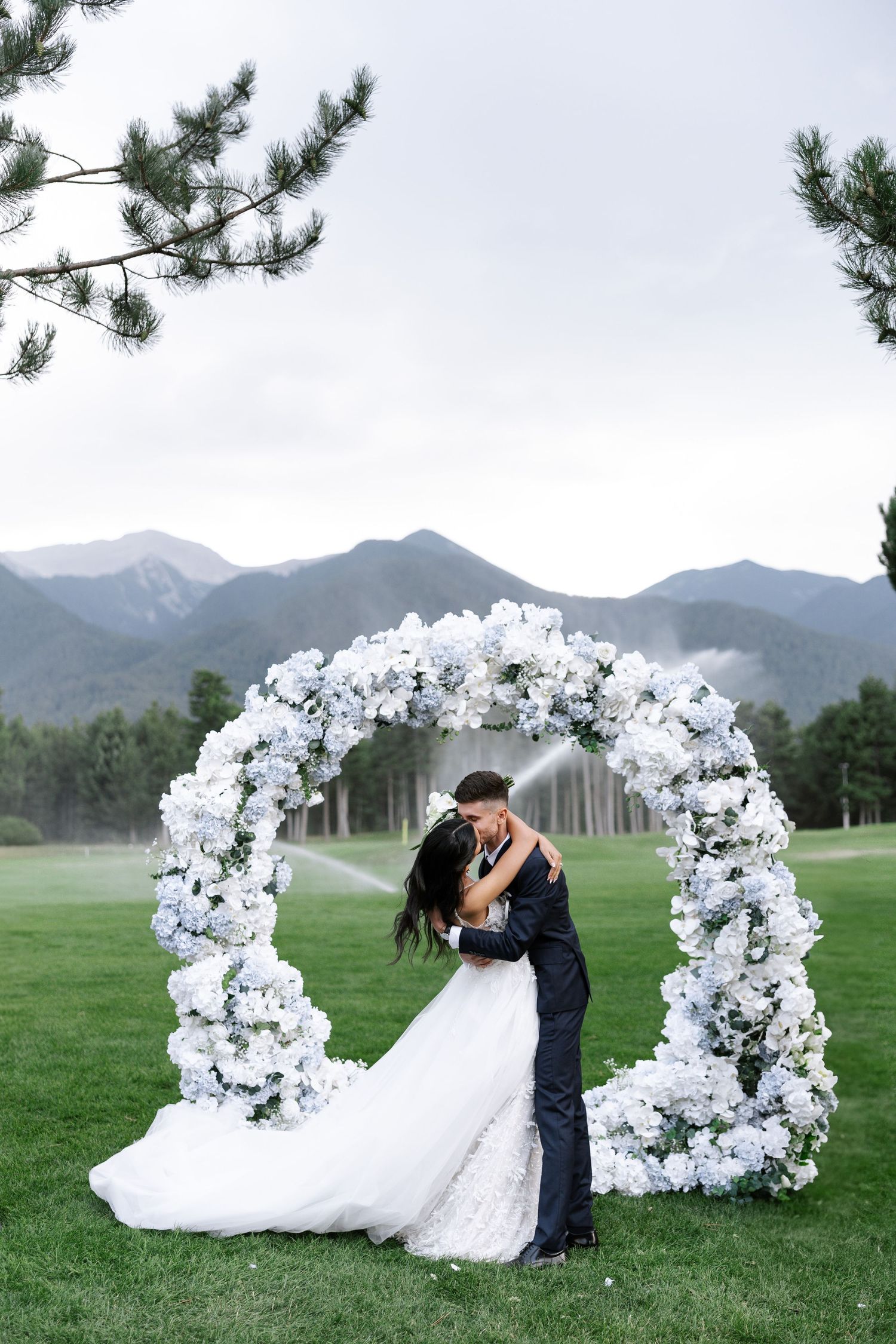 A romantic wedding scene with a floral arch of white blooms set against mountain views on a lush green lawn.