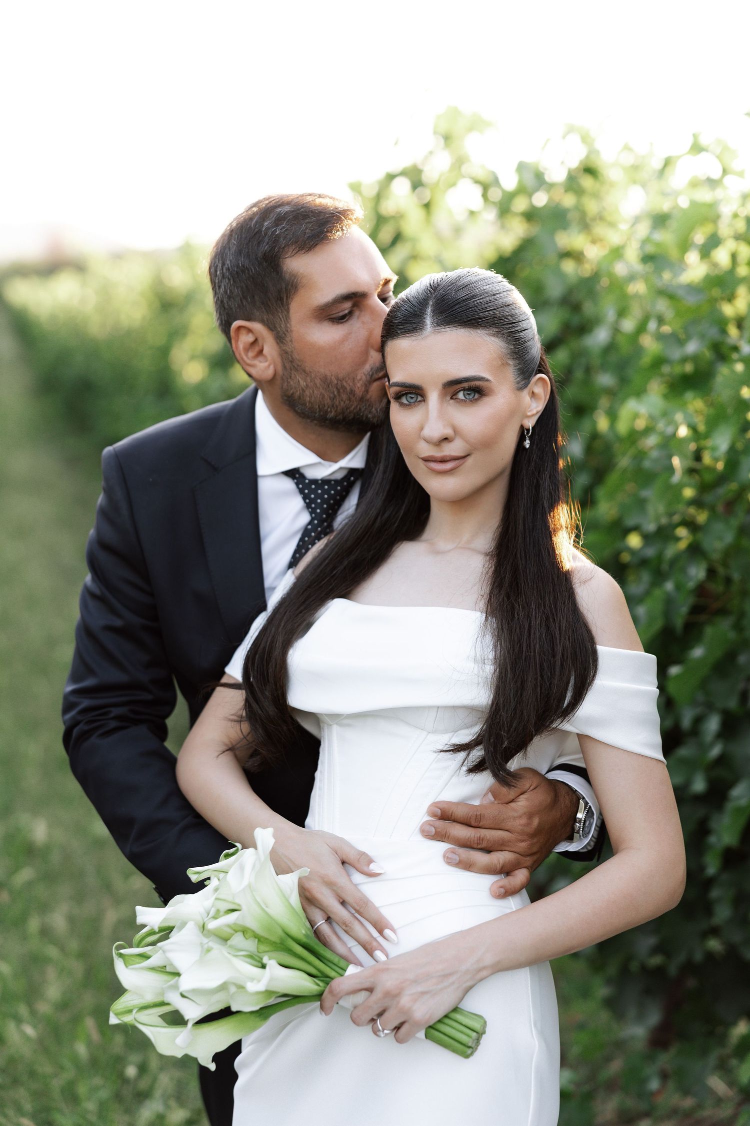 A romantic wedding couple embraces in a vineyard setting with white calla lily bouquet at sunset.