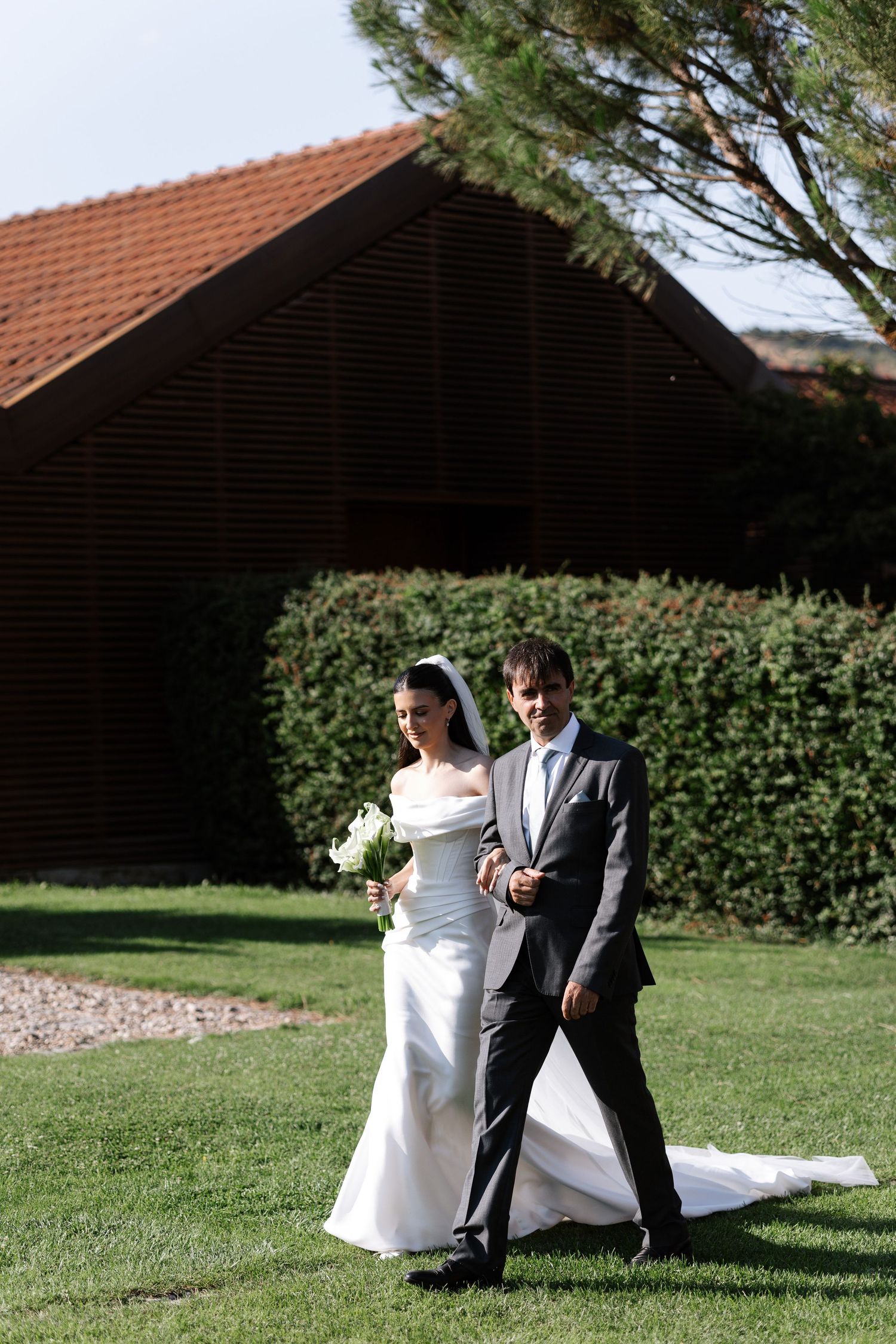 A couple in wedding attire playfully poses together on a sunny lawn with green hedges and buildings in the background.