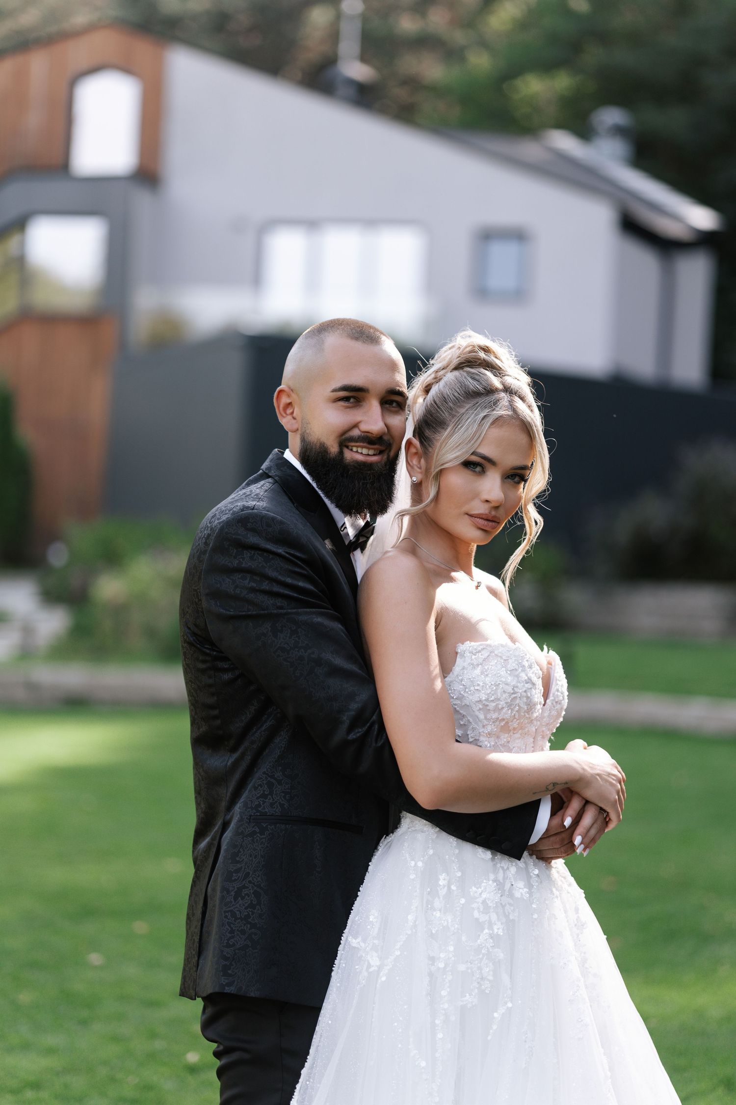 A couple shares romantic moments in formal wedding attire while posing together outdoors on a lawn.