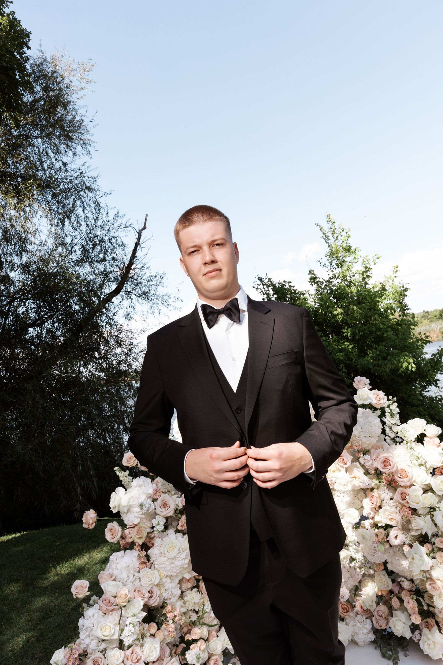 Formal portrait of a groom in black tuxedo standing by floral arch with pink and white roses outdoors.