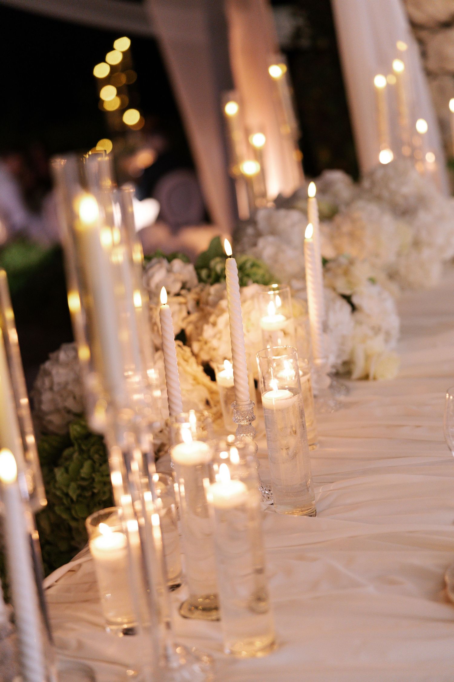 Elegant wedding reception table decorated with glowing white candles and floral arrangements on a white tablecloth.