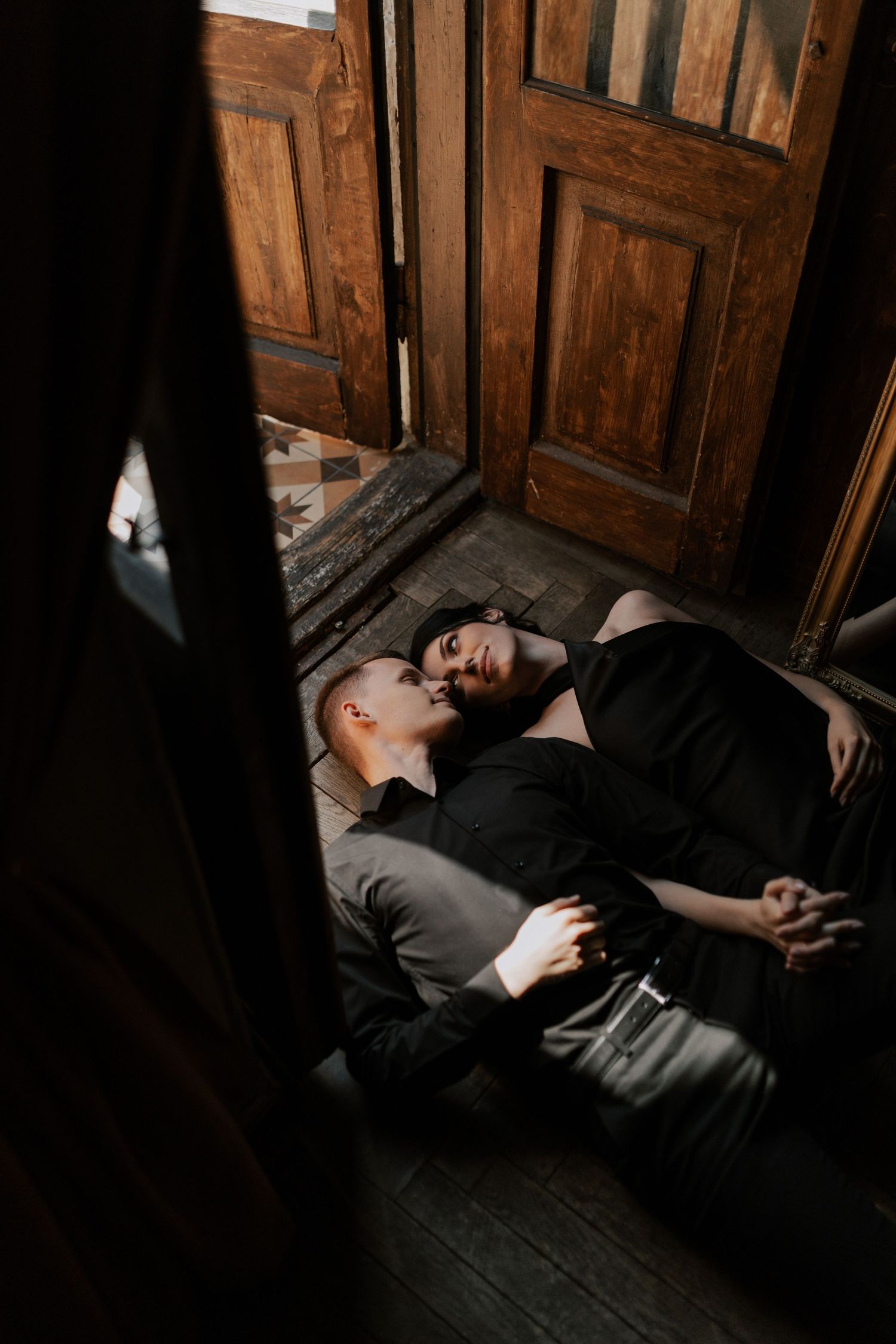 Intimate couple lying on wooden floor near vintage doors in dramatic lighting.