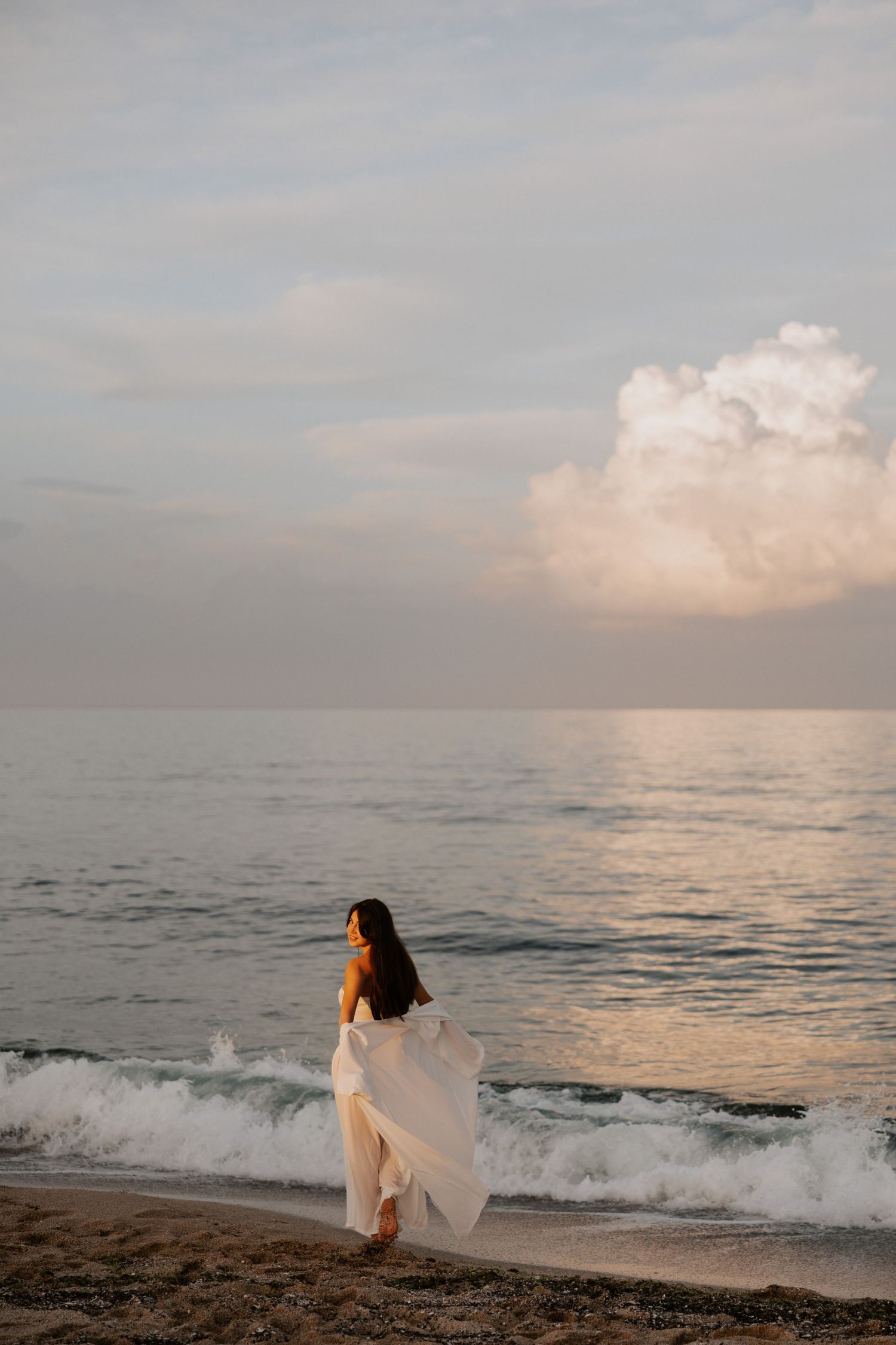 A woman in a flowing white dress stands by the ocean during sunset.