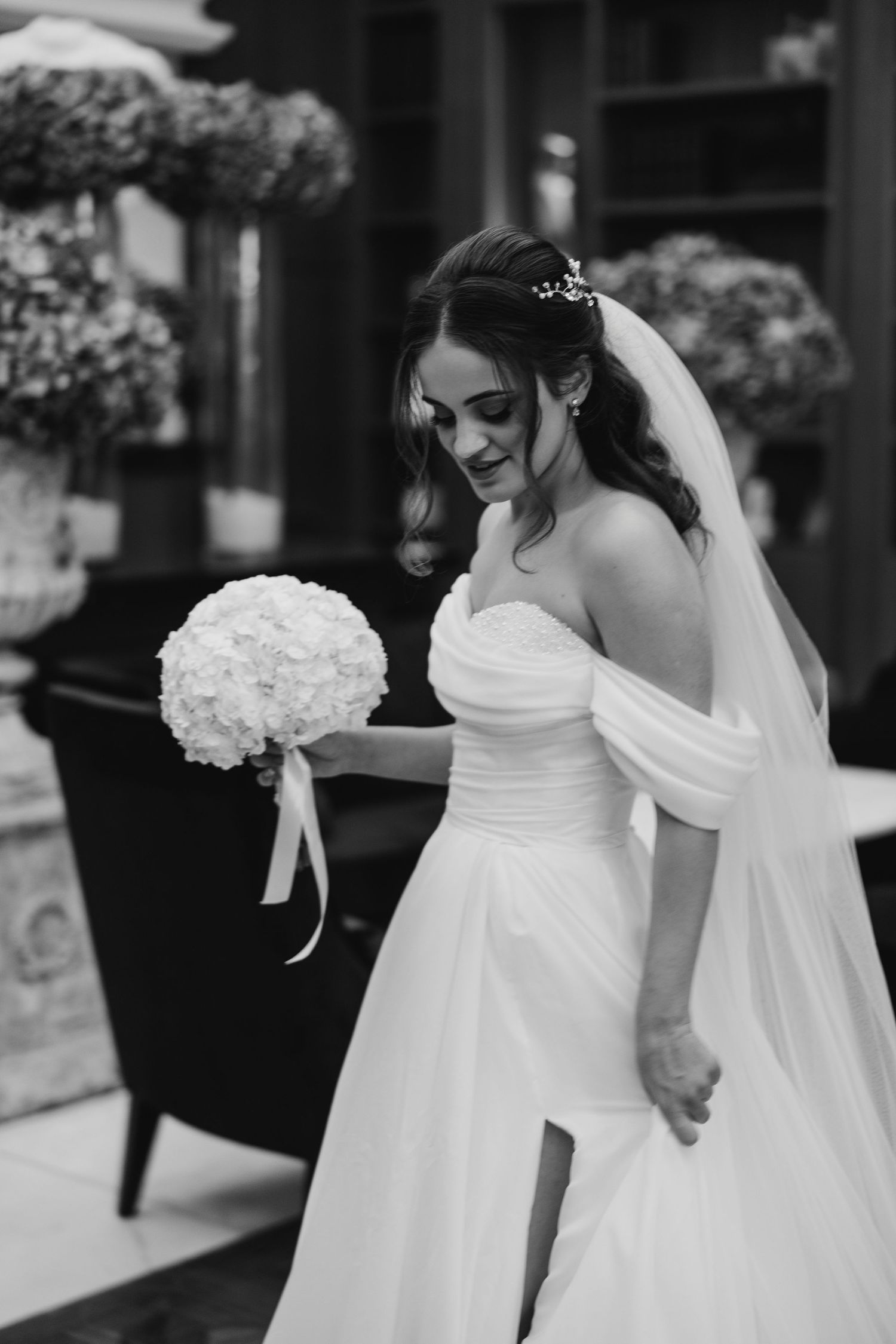 Bride in a flowing white off-shoulder wedding gown holds a white hydrangea bouquet in elegant black and white.