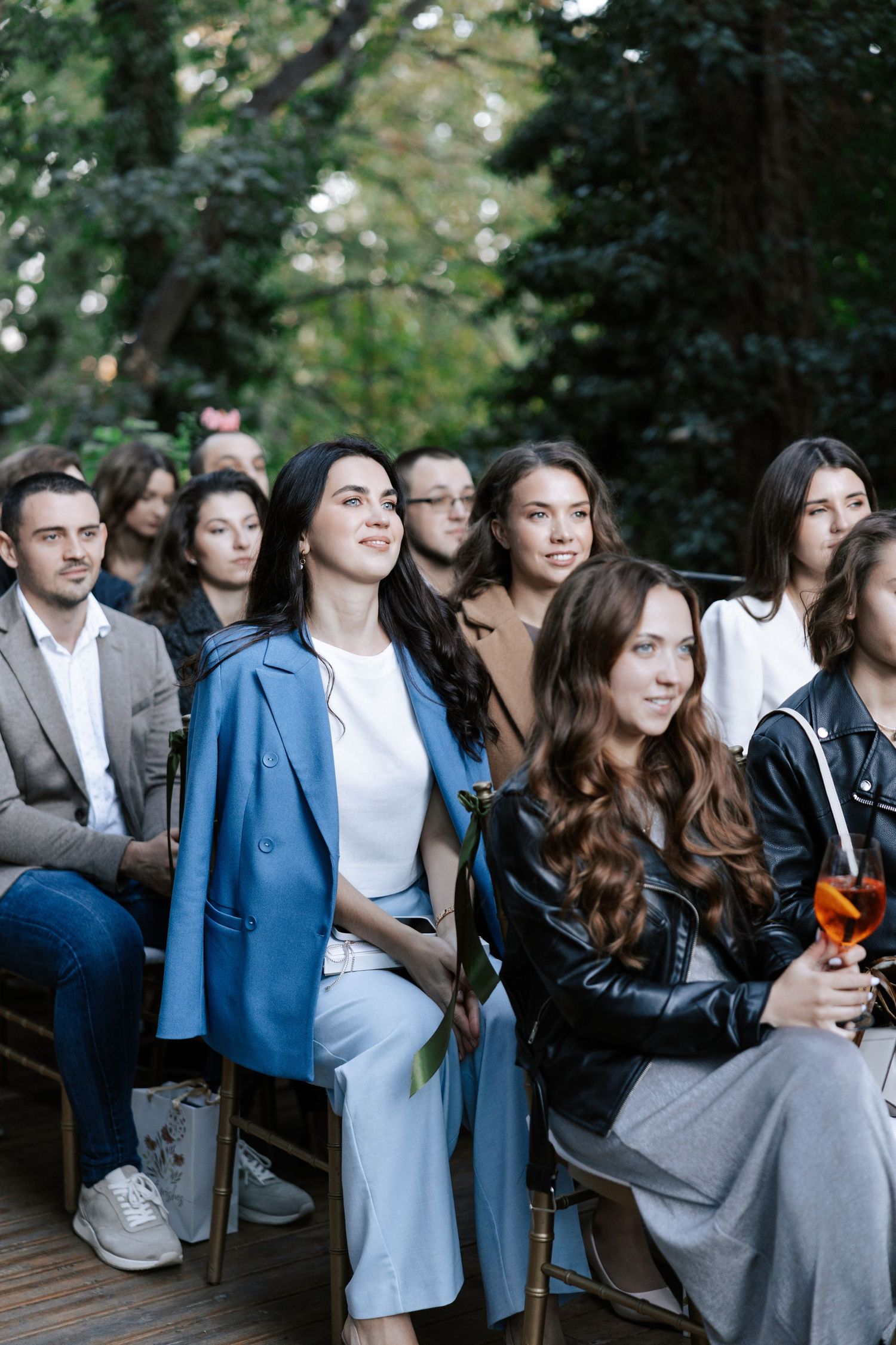 Wedding guests are seated outdoors at a ceremony venue with lush greenery in the background.