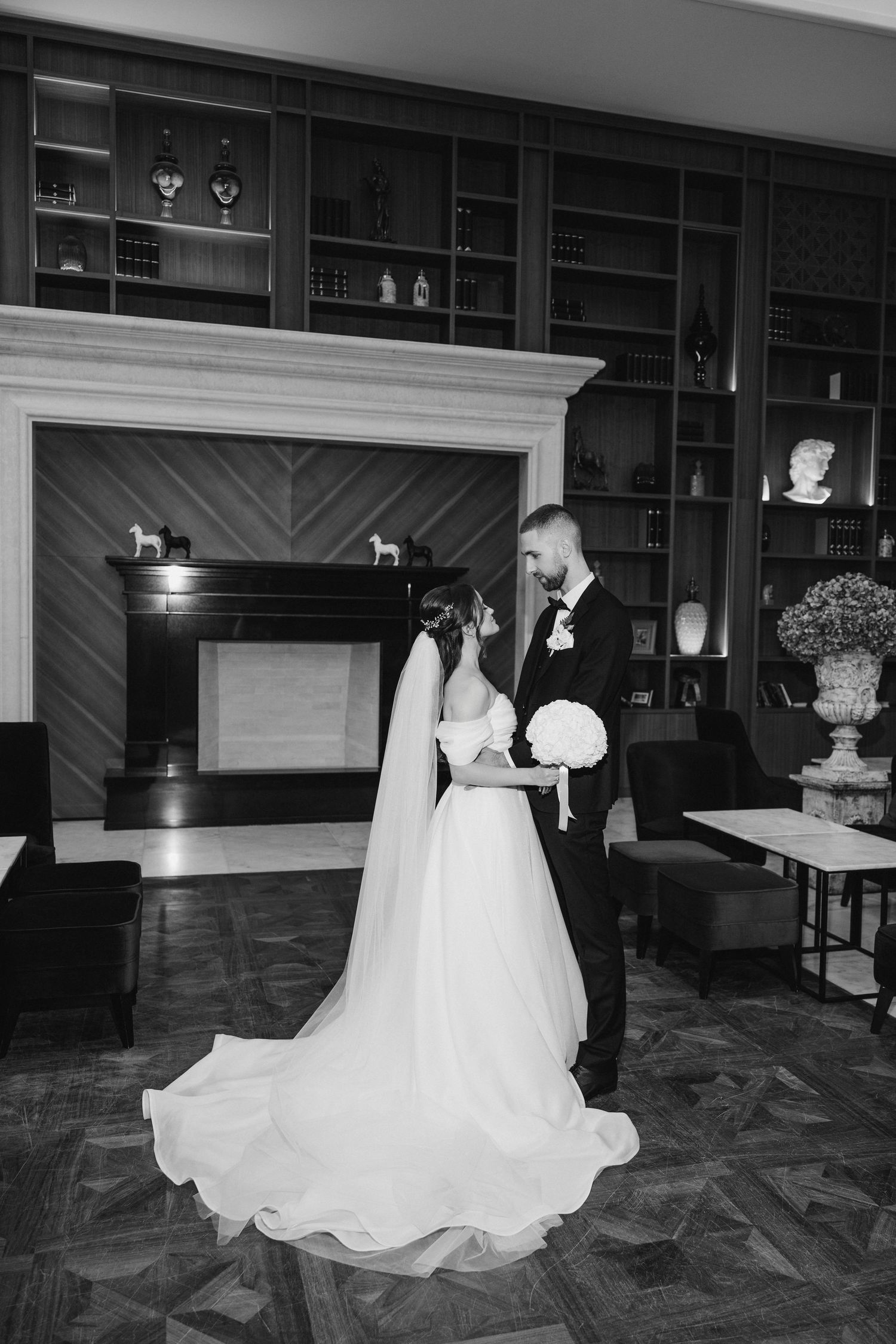 Black and white photo of wedding couple posing in front of an elegant fireplace with built-in shelving.