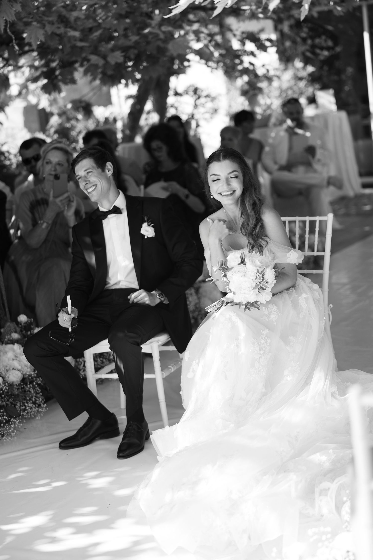 A bride and groom share a joyful laugh while seated at their outdoor wedding ceremony under dappled sunlight.