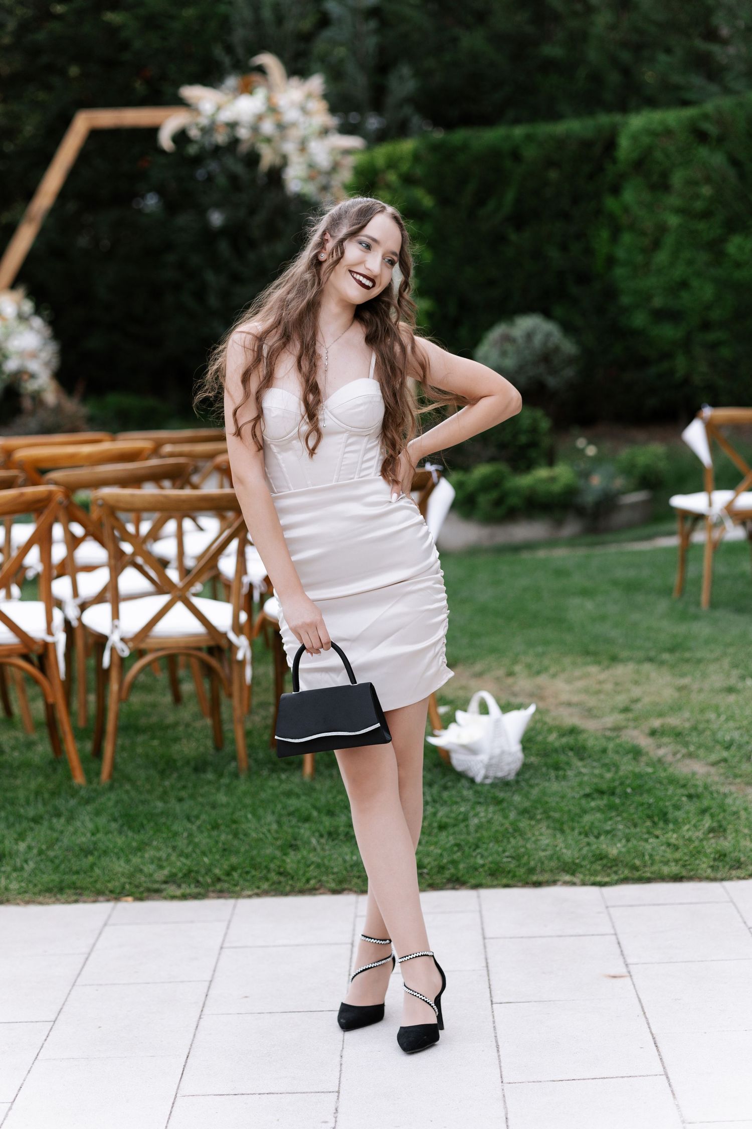 A woman in a white dress and black heels poses on stone pavement with wooden chairs in the background.