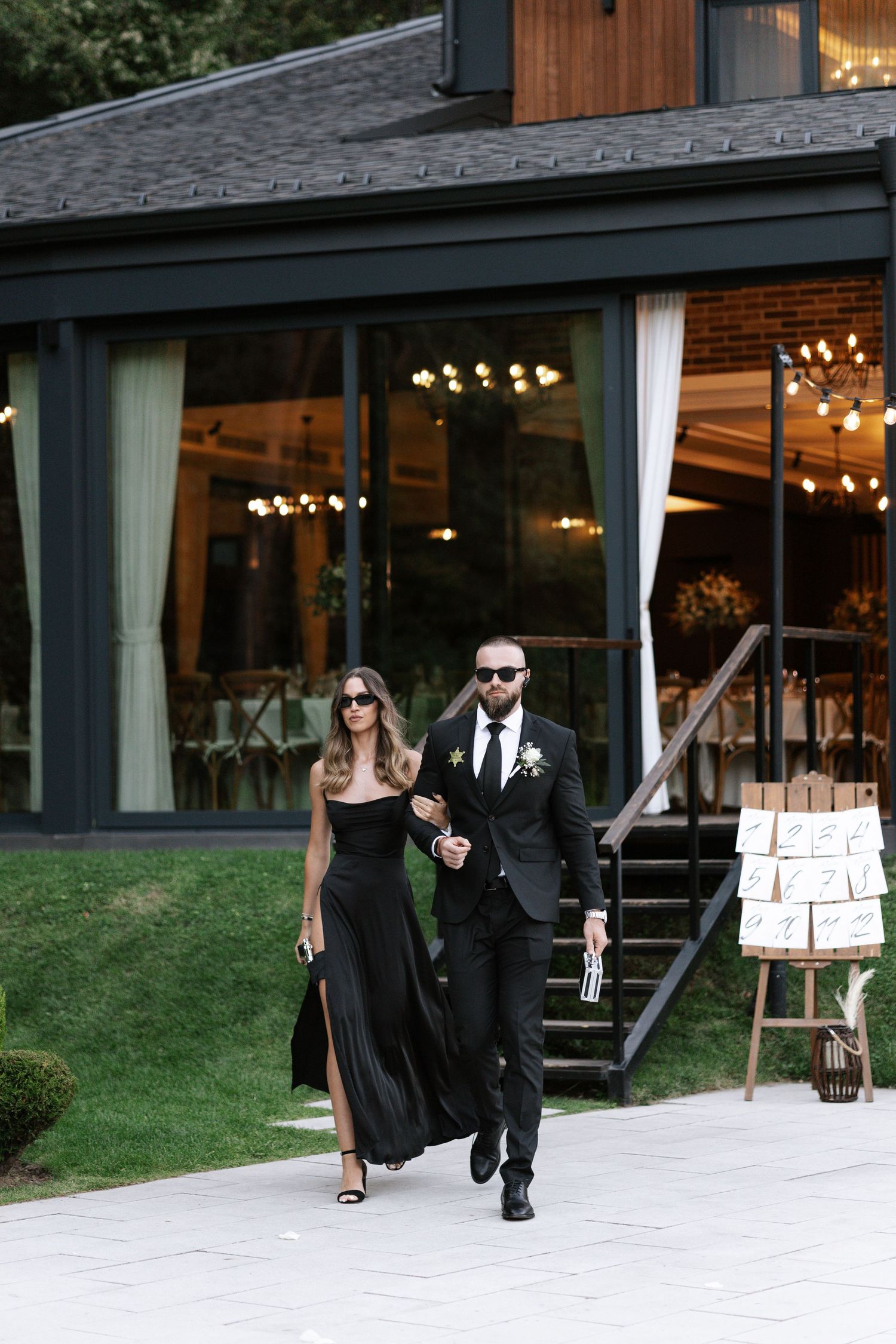 A couple in formal black attire walks together on a concrete path outside a modern building with glass windows.