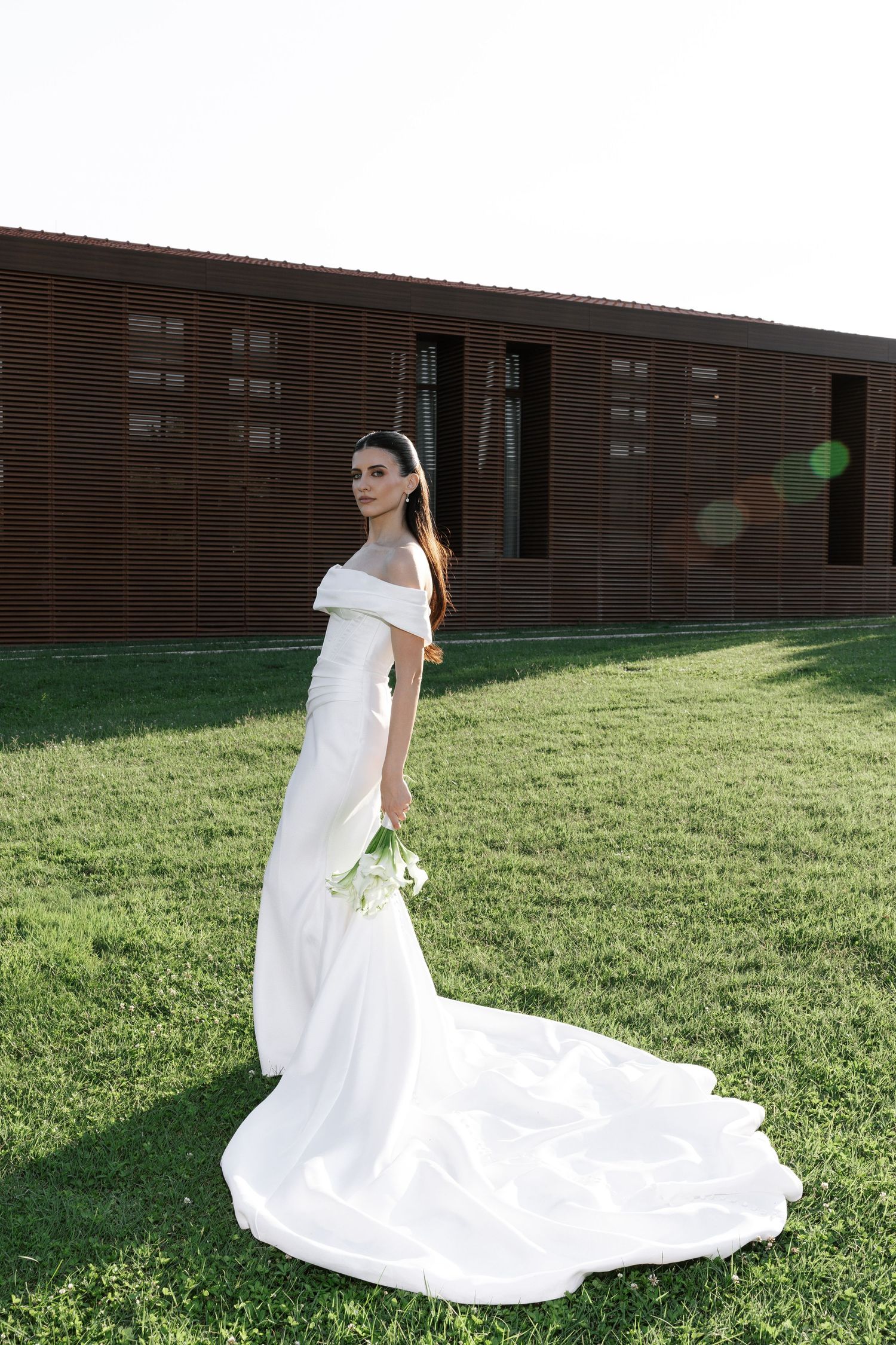 A bride in a classic white wedding dress poses gracefully on a grassy lawn beside a brick building.