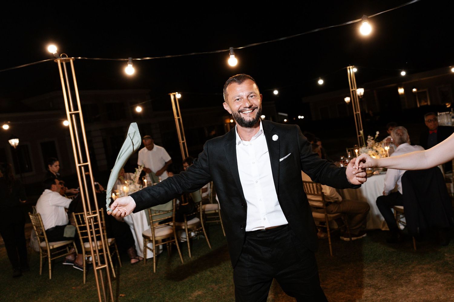 String lights illuminate an outdoor evening wedding reception with guests seated at tables under a dark sky.