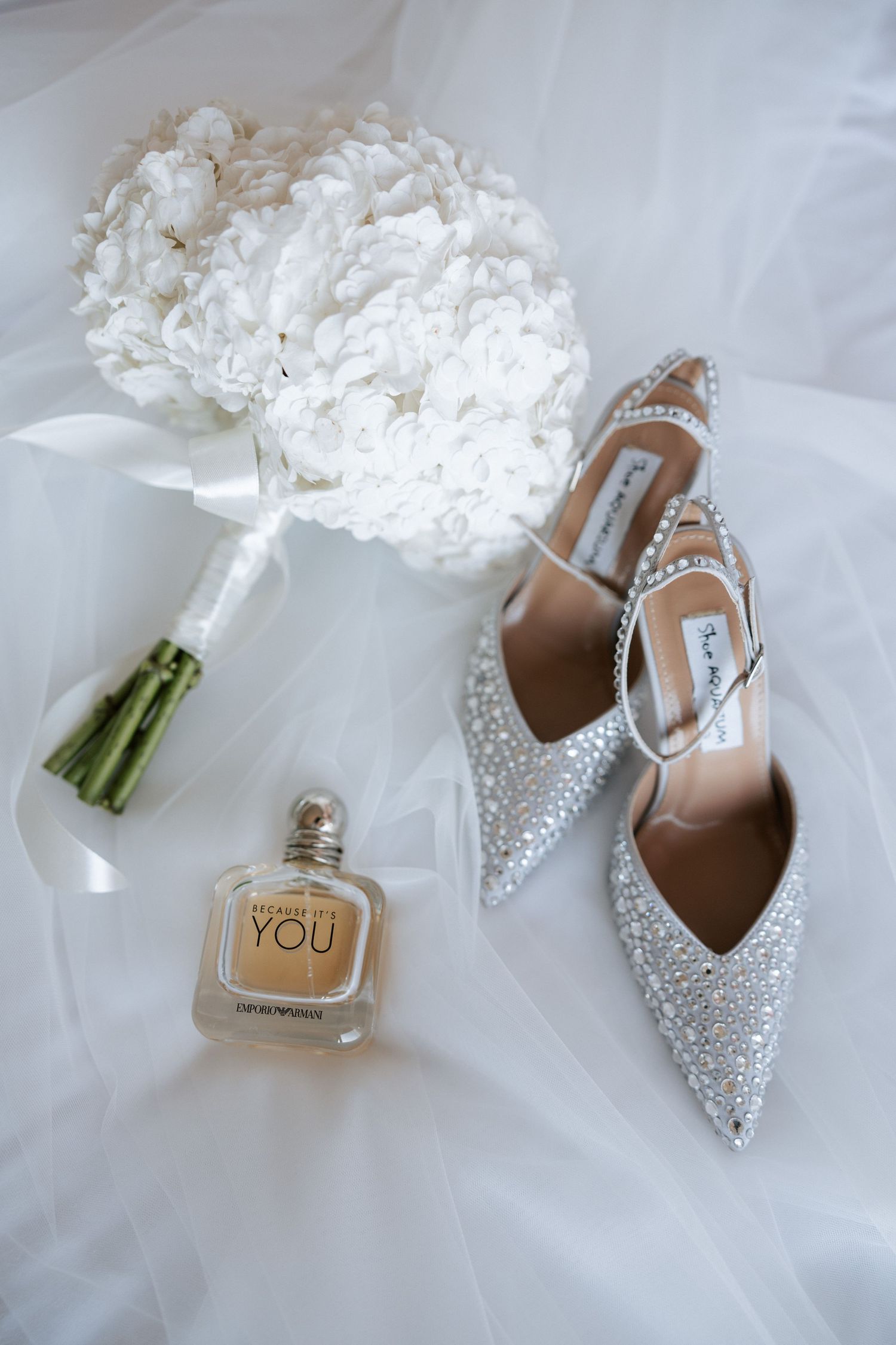 Wedding accessories including crystal shoes, white bouquet and perfume bottle arranged on white tulle.