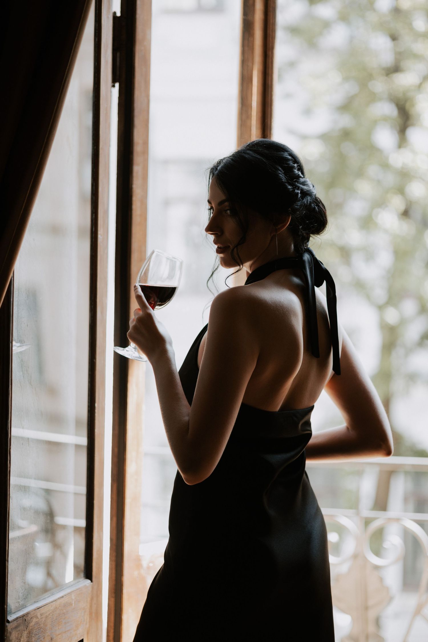 Silhouette of a woman in a black dress holding a wine glass by a window.