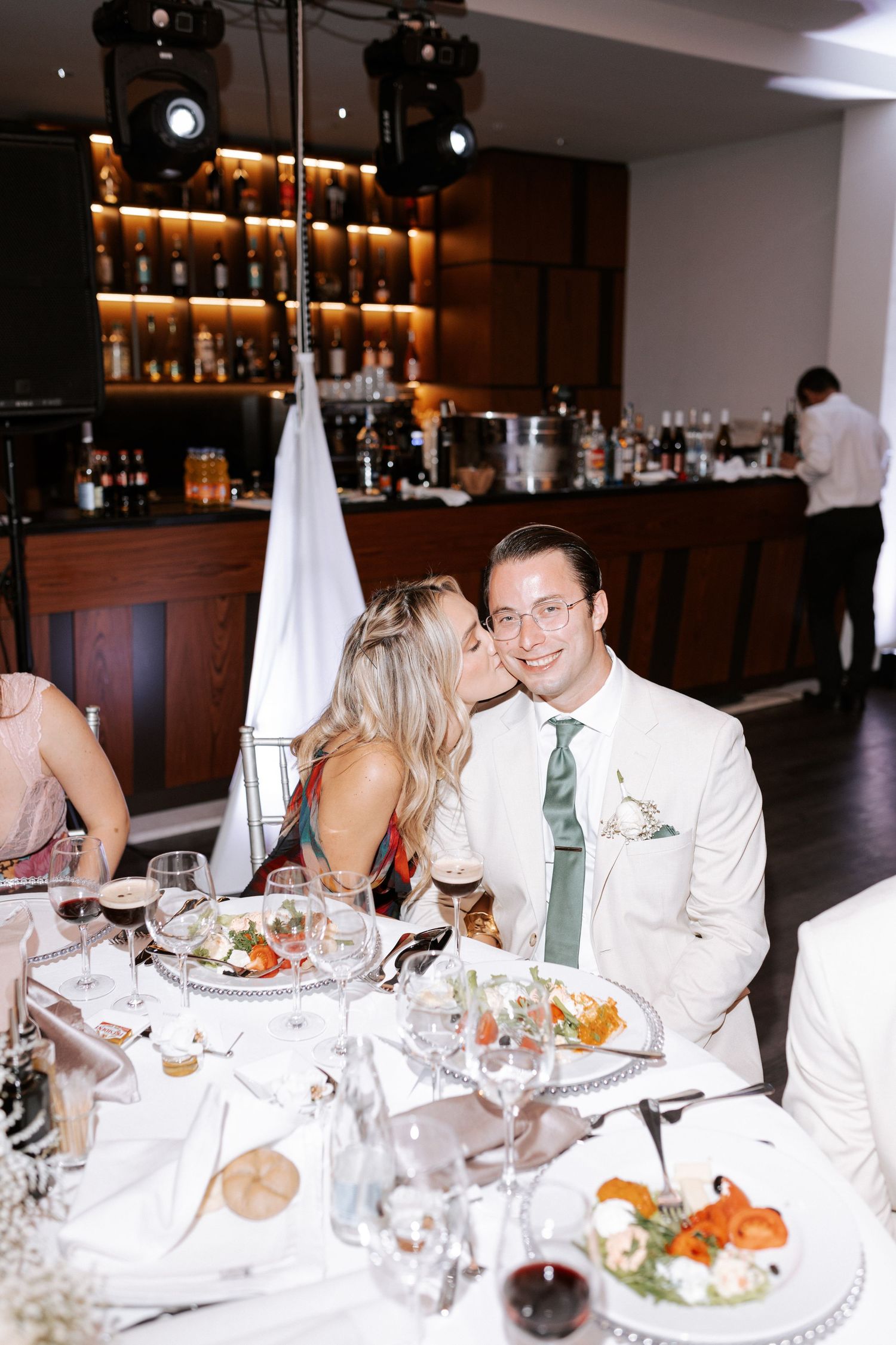 Wedding guests enjoy dinner at an elegant reception with bar in background.
