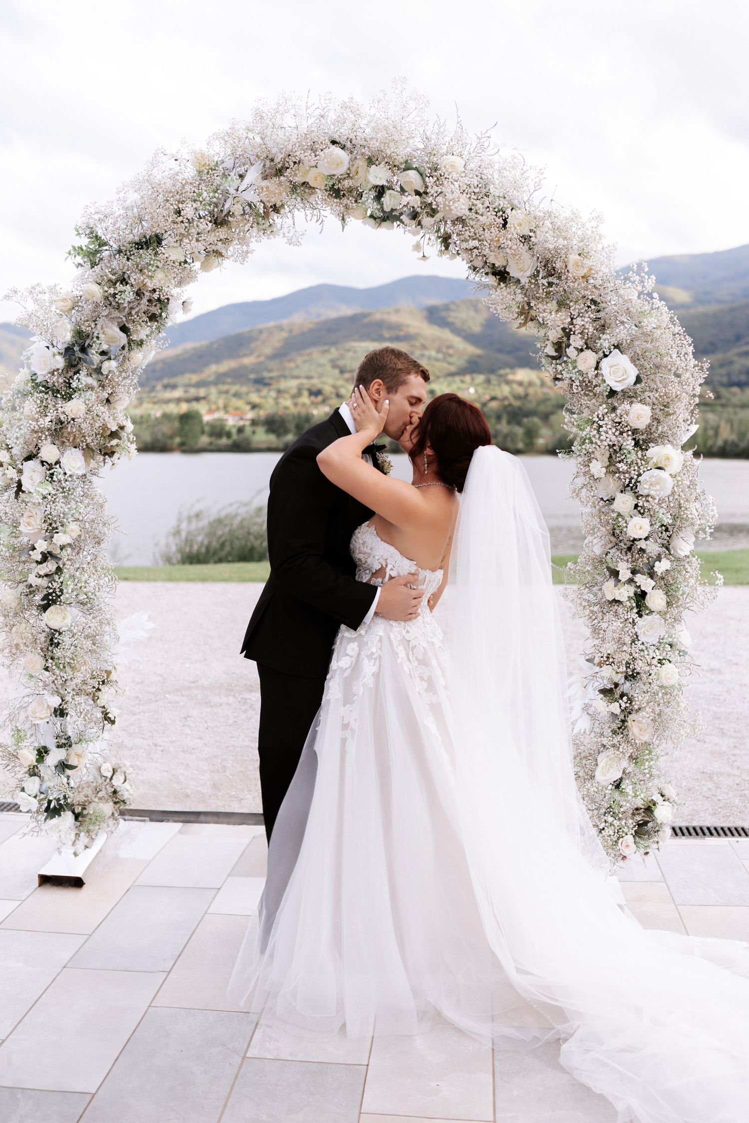 Romantic moment under white floral arch with mountain and lake views.