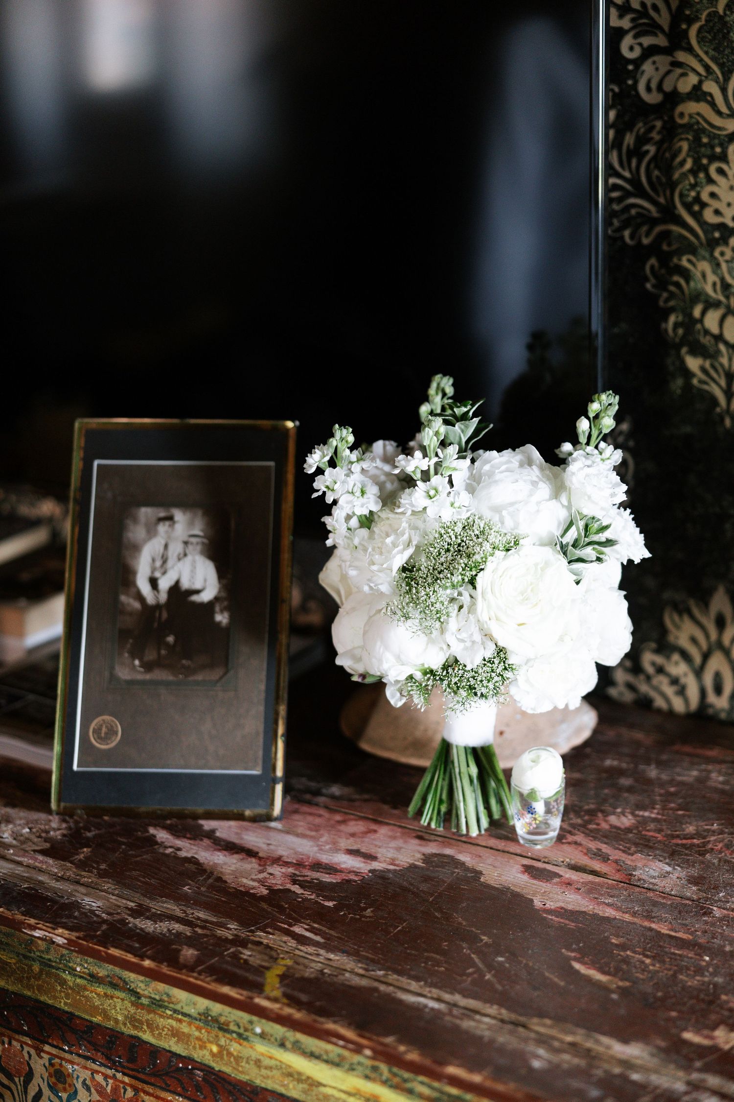 A vintage photograph frame sits beside an elegant white floral bouquet with greenery on a rustic wooden surface.