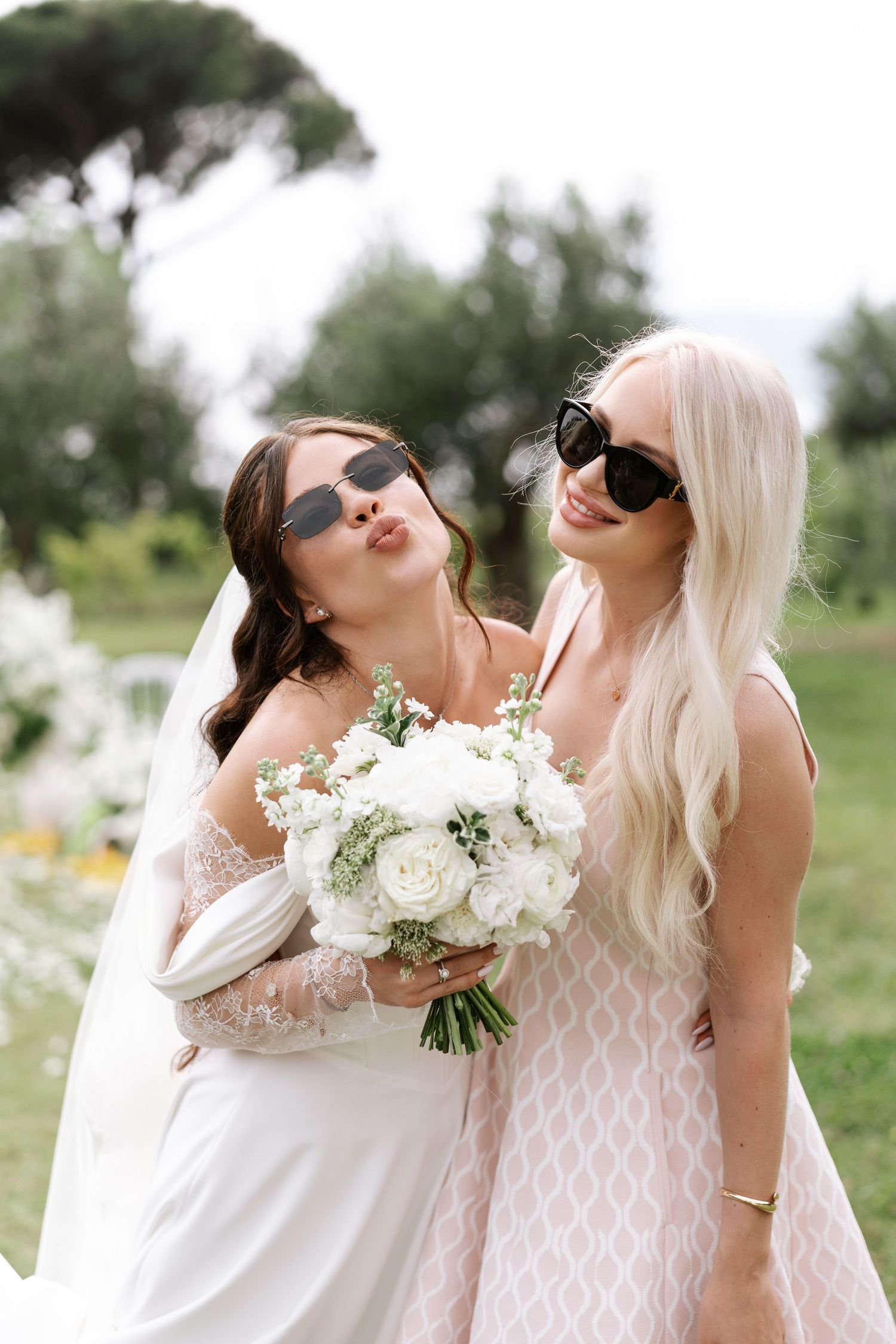 Two people in white dresses and sunglasses pose playfully with a white floral bouquet in an outdoor garden setting.