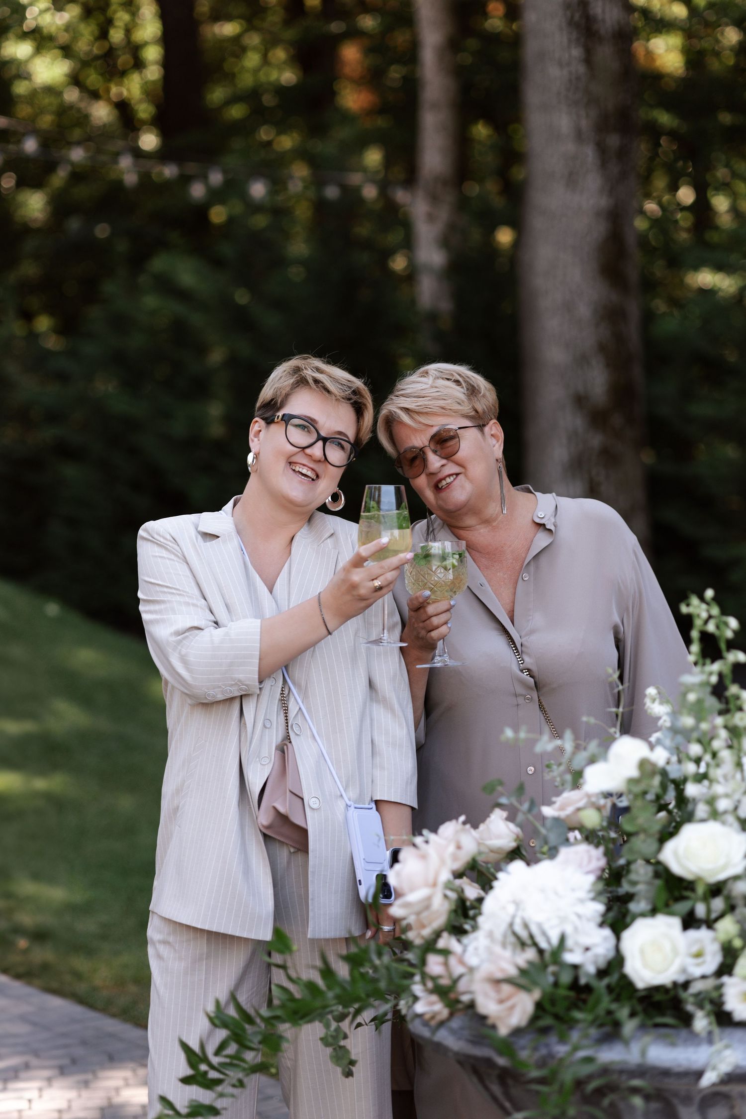 Two people toast with glasses while surrounded by white floral arrangements in an outdoor garden setting.