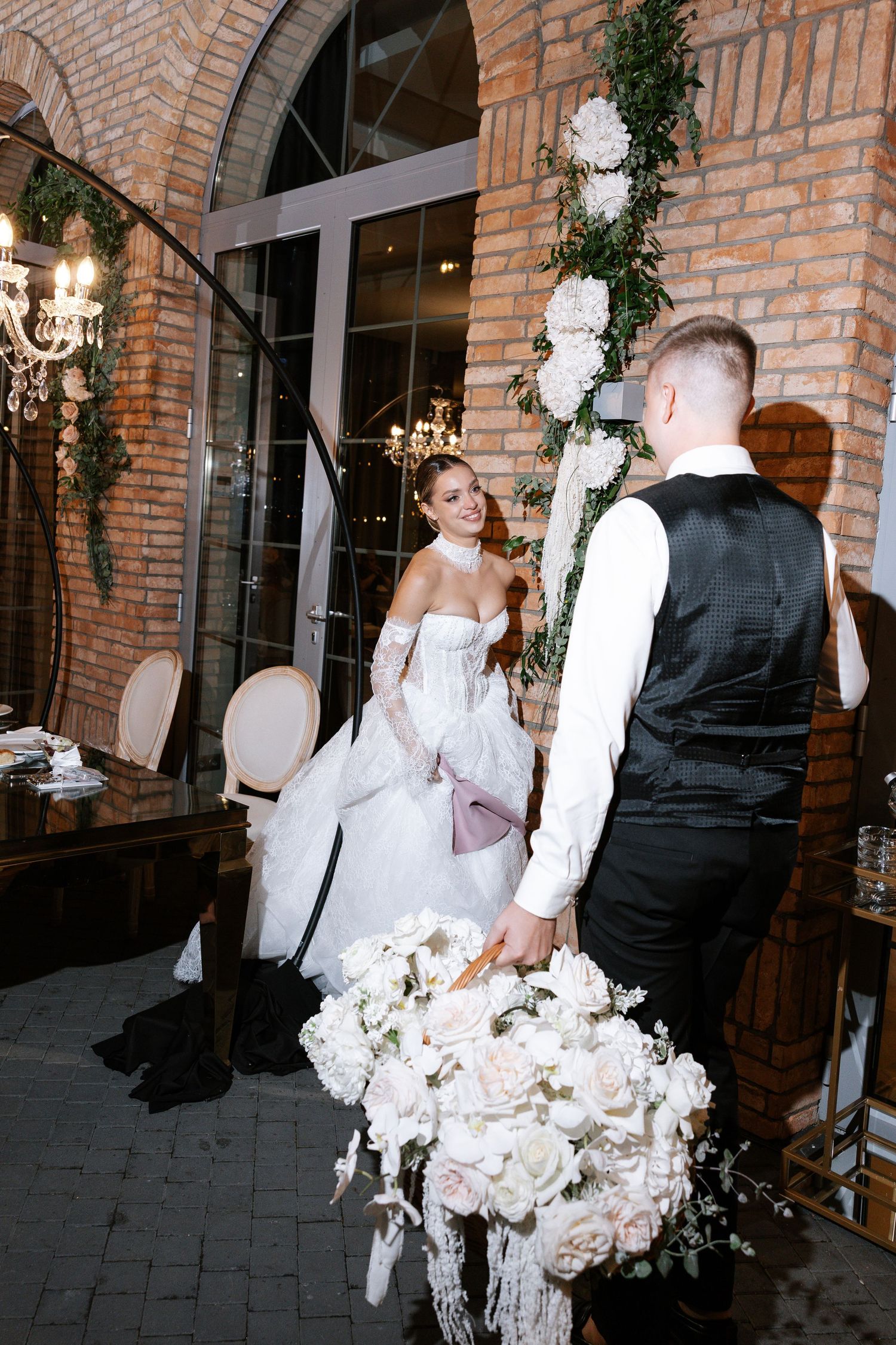 Candid wedding moment captured near brick archway decorated with white floral arrangements.