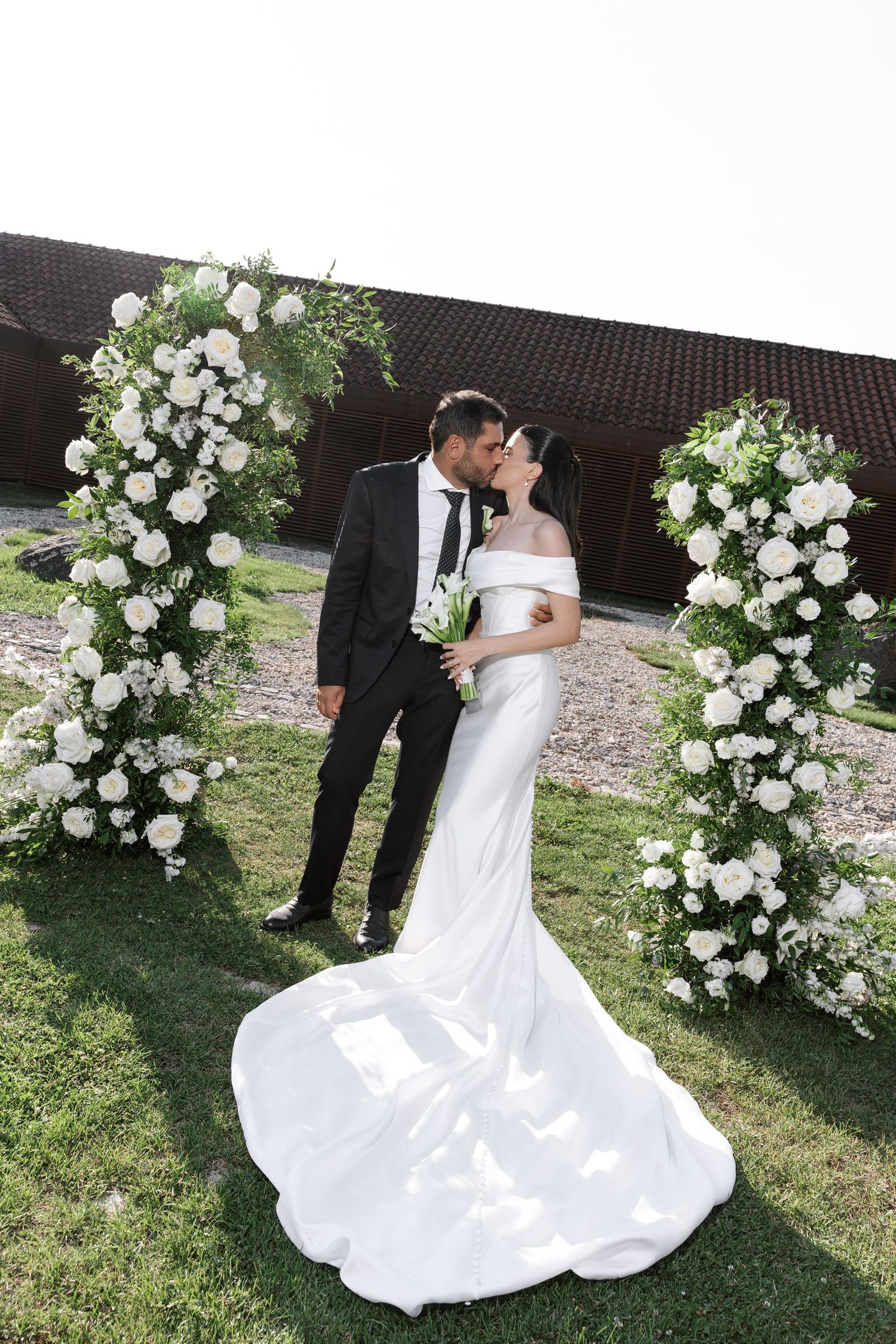 A romantic wedding couple poses under a beautiful white floral arch in an outdoor garden setting.