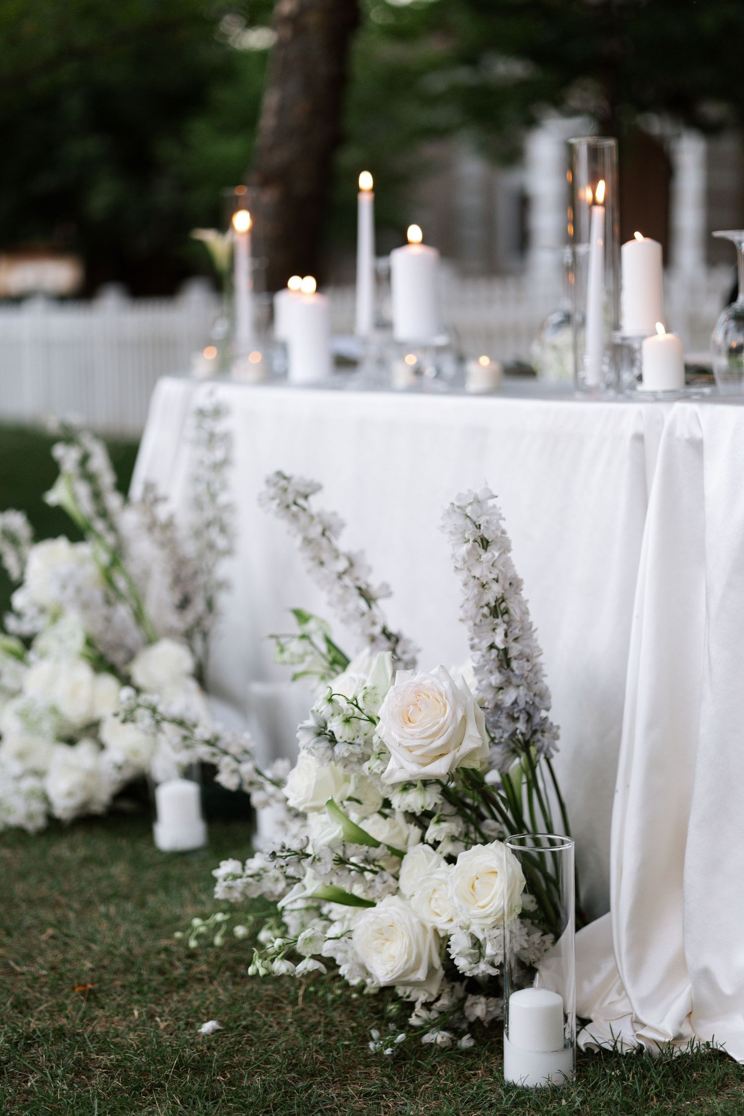 White taper candles and elegant floral arrangements with roses and baby's breath line a romantic outdoor wedding setting.