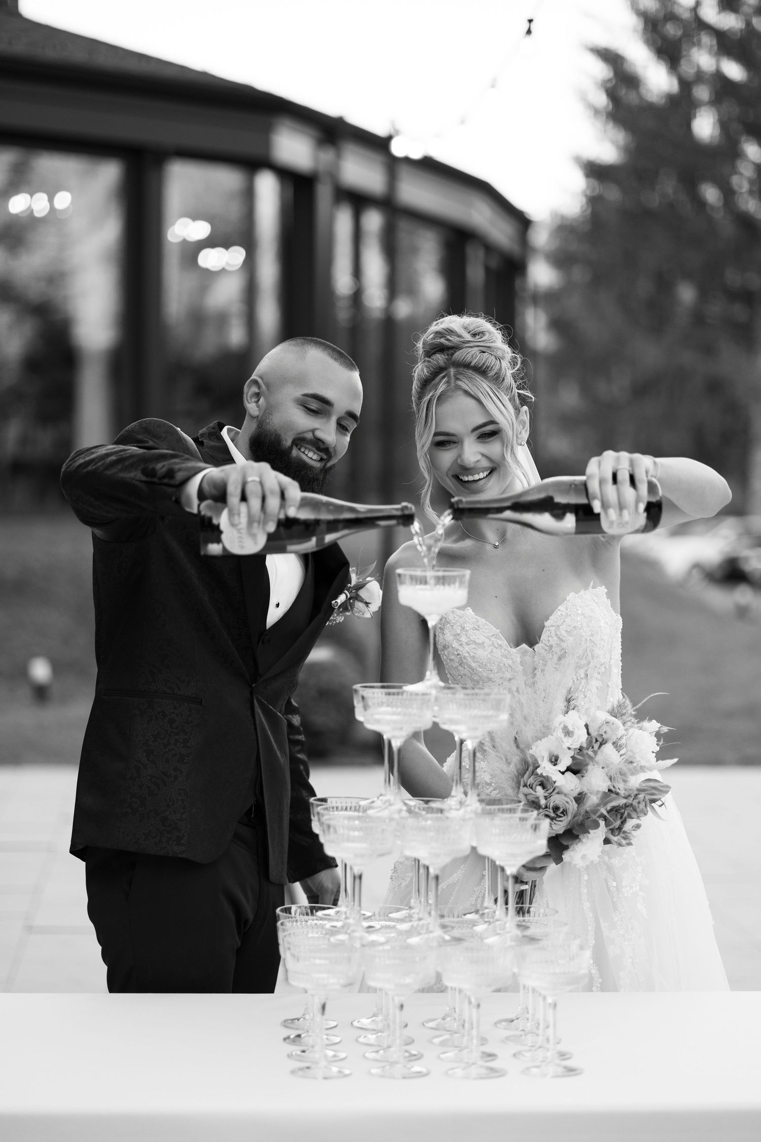 Wedding couple pours champagne into tower of crystal glasses during elegant reception celebration.