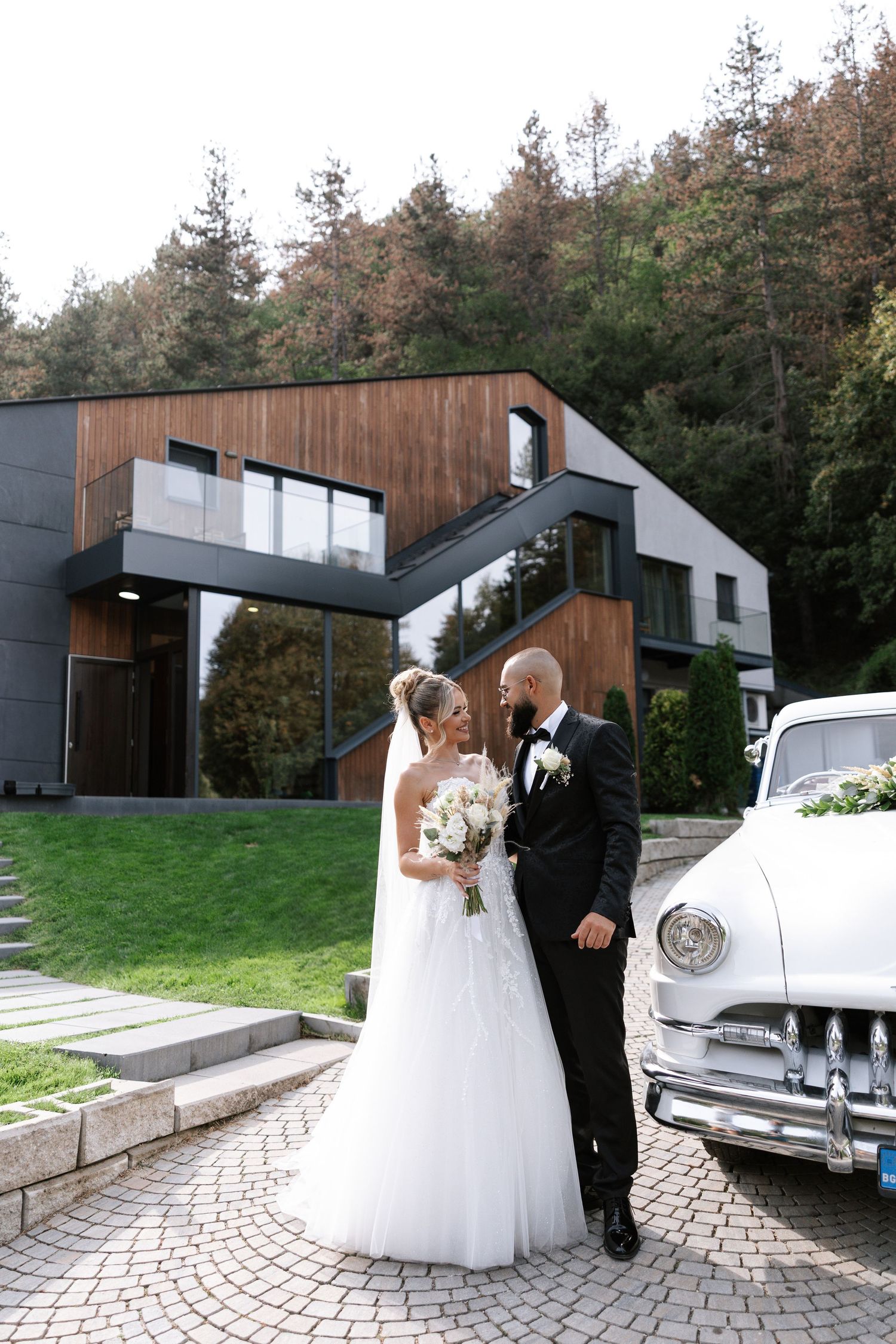 A wedding couple poses beside a classic white vintage car in front of a modern architectural venue with wooden accents.