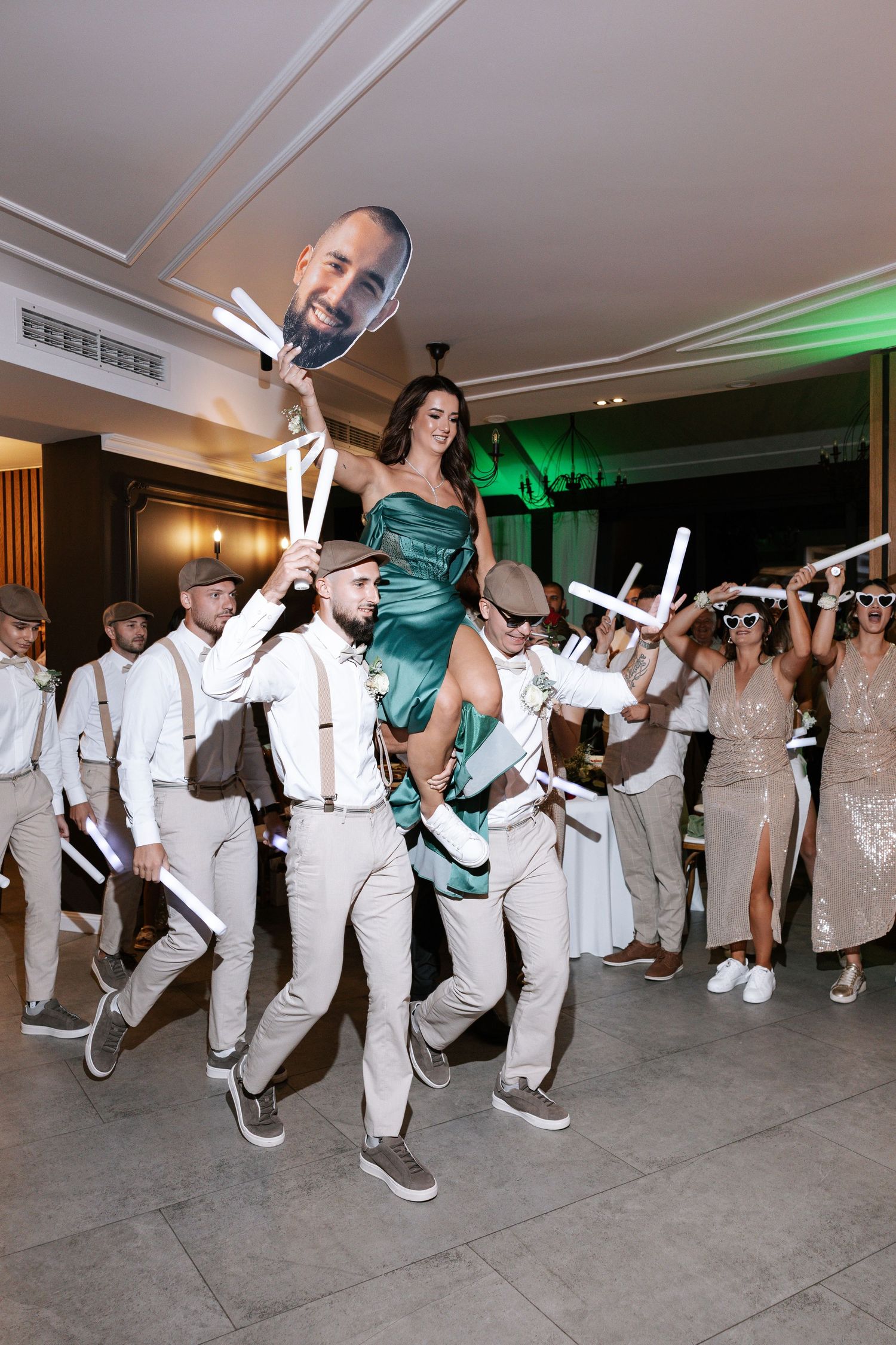 Wedding party in white attire performs synchronized dance moves in a reception hall with green accent lighting.