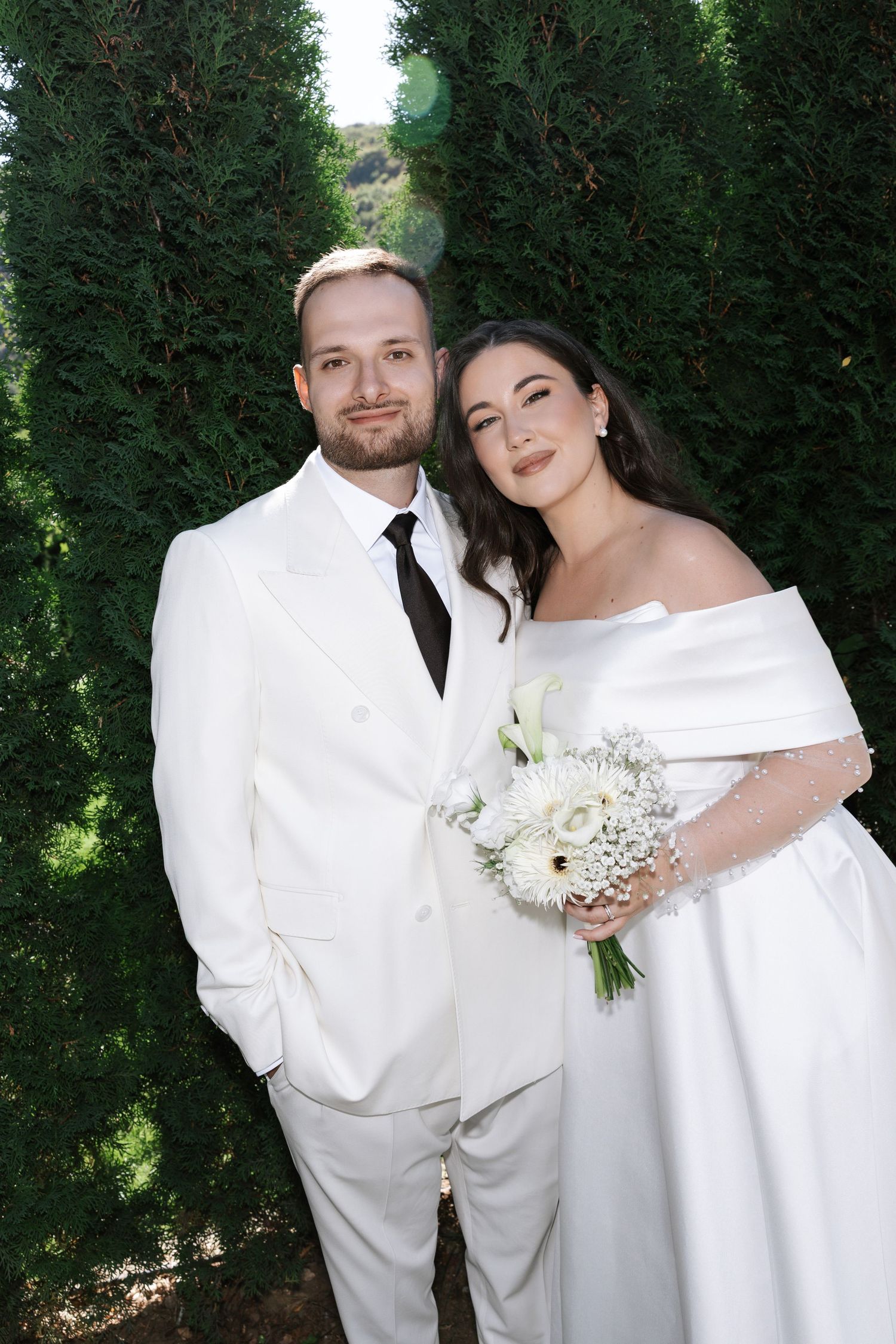 Newlywed couple in elegant white attire poses for romantic outdoor wedding photos against lush green hedges.