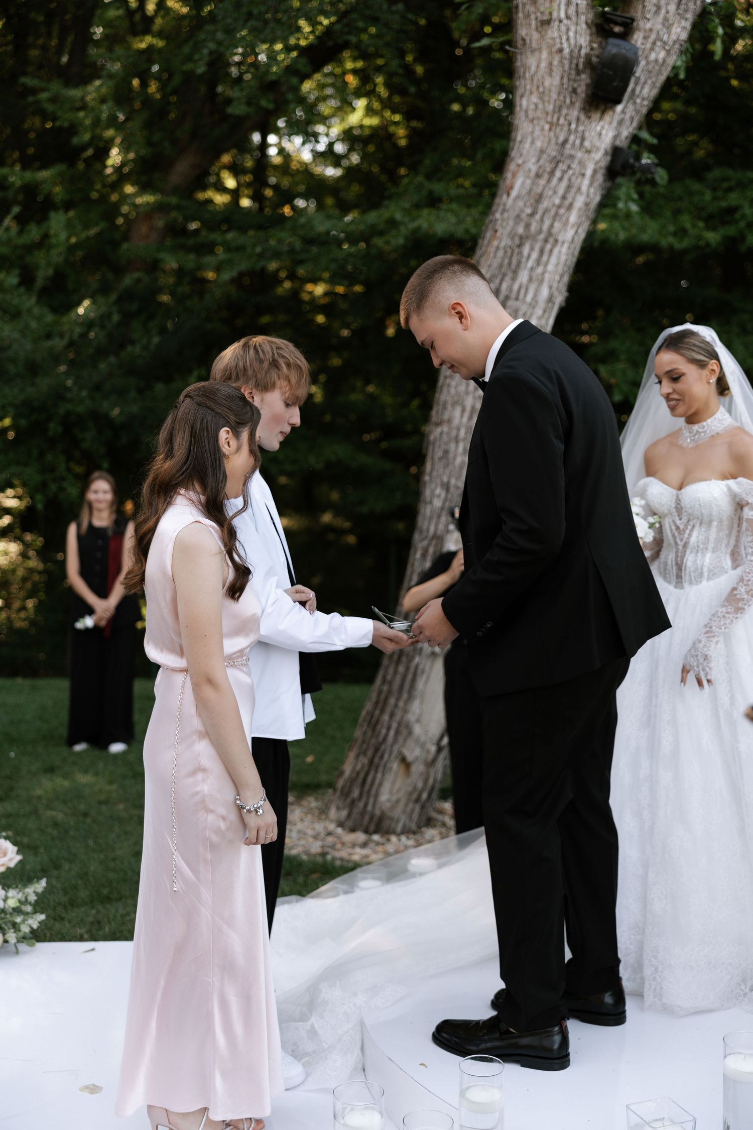 Wedding ceremony taking place outdoors with attendees gathered around a tree on a white aisle runner.