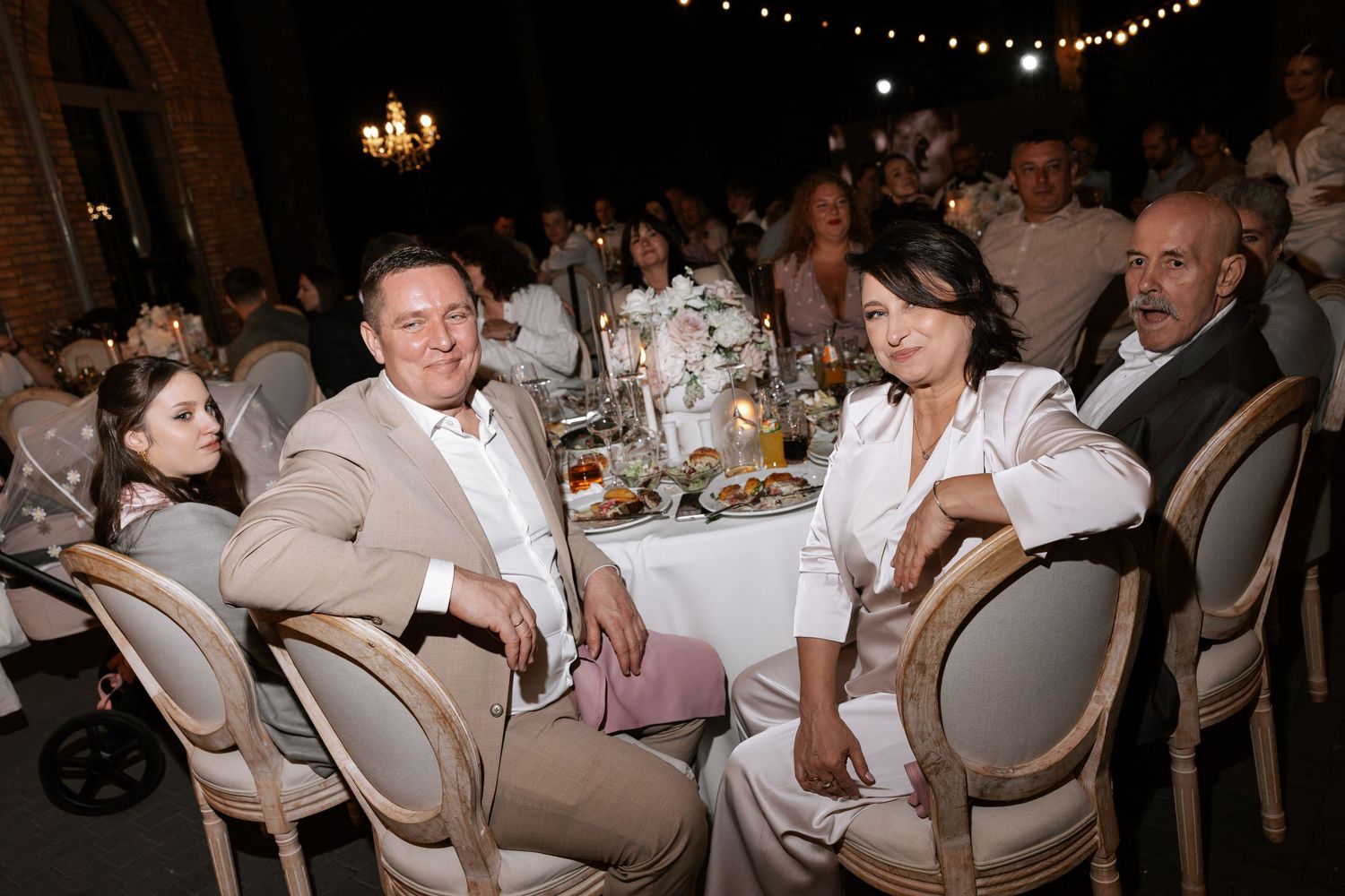 Wedding guests enjoy dinner at an elegant reception with string lights and chandelier decor.