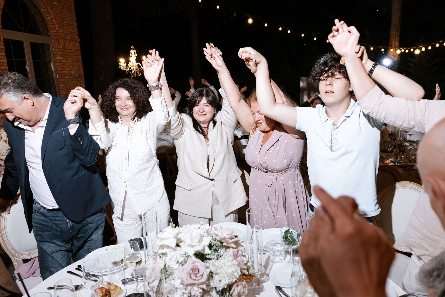 Wedding guests raise their hands in celebration during an evening reception.