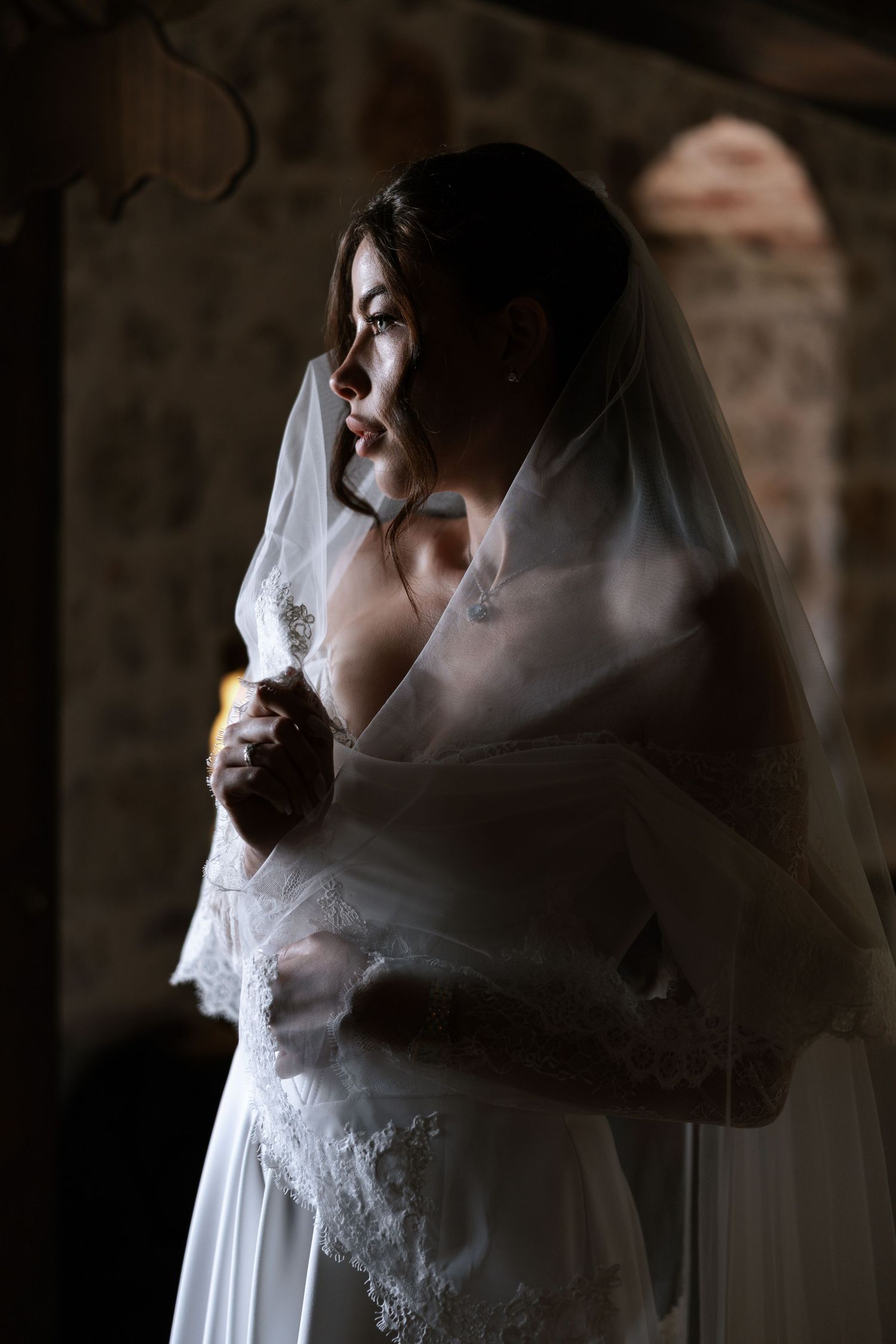 A bride in white veil poses in dramatic side lighting against a dark background creating an ethereal silhouette.