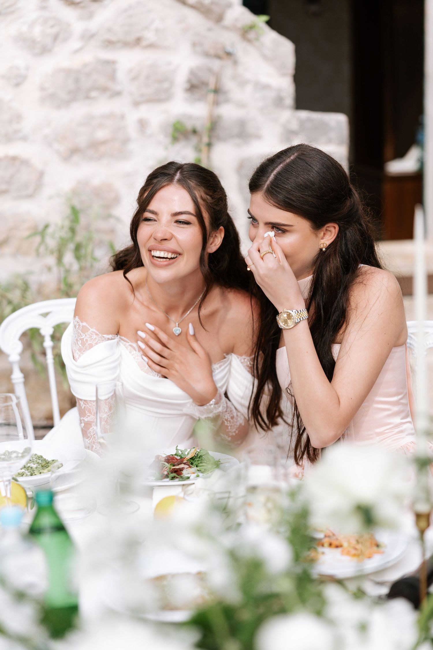 Two women in white and pink dresses share a joyful moment at a wedding reception table.