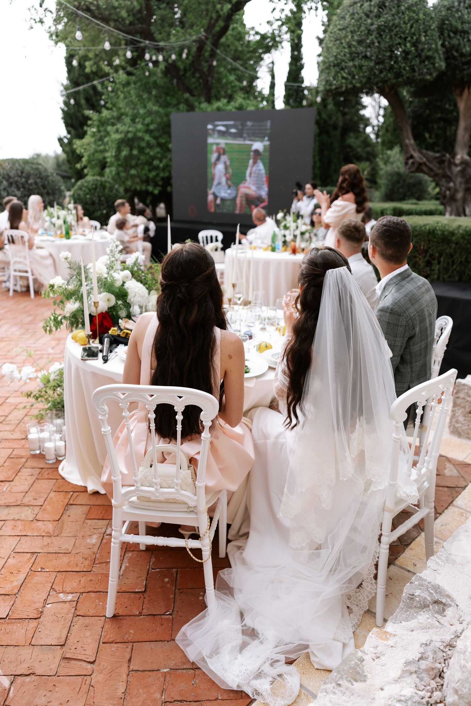 Wedding reception guests seated at elegant outdoor tables with white decor and flowing veil details on brick patio.