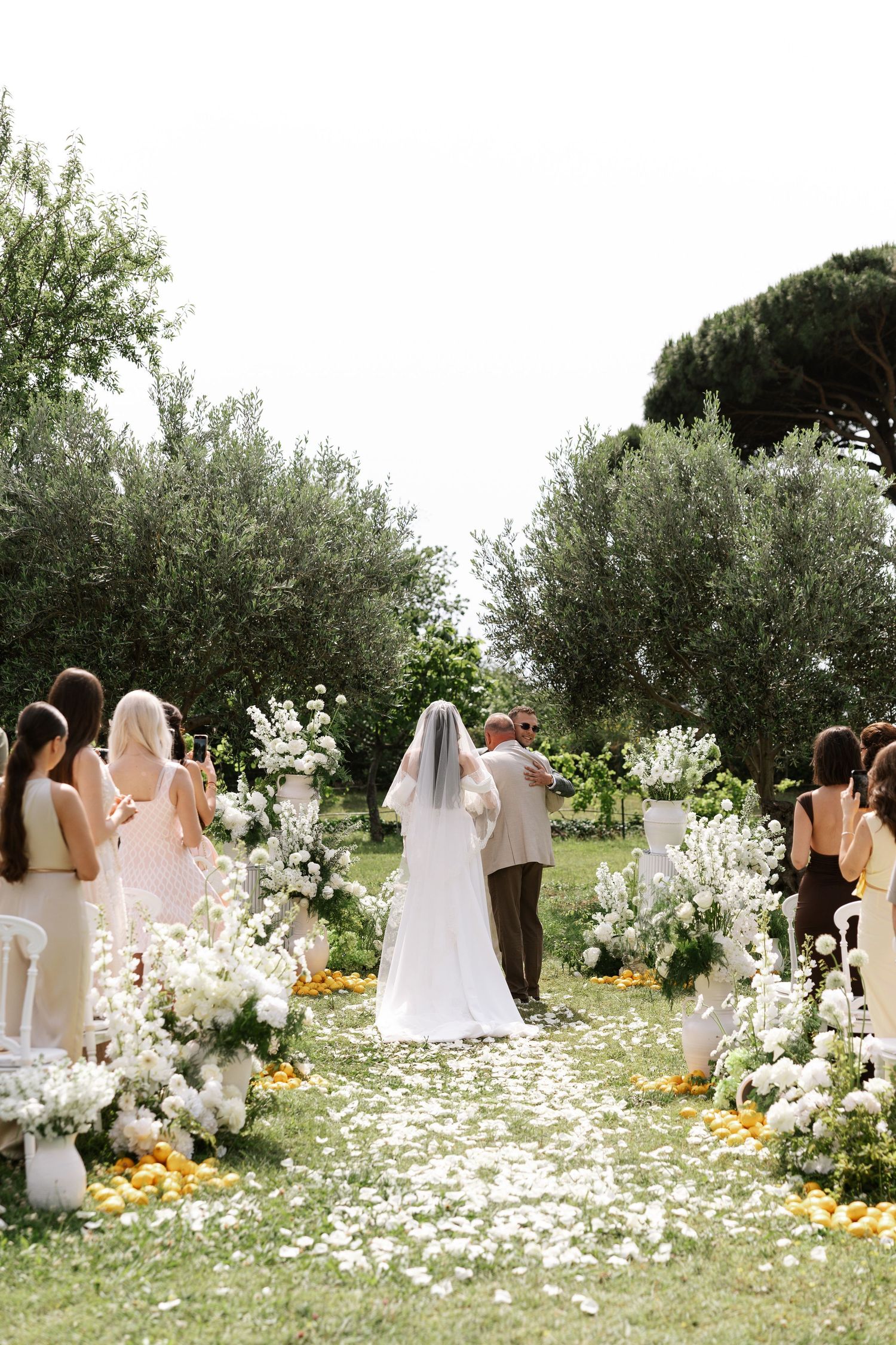 A wedding ceremony setup in an olive grove with white floral arrangements and scattered petals on the ground.