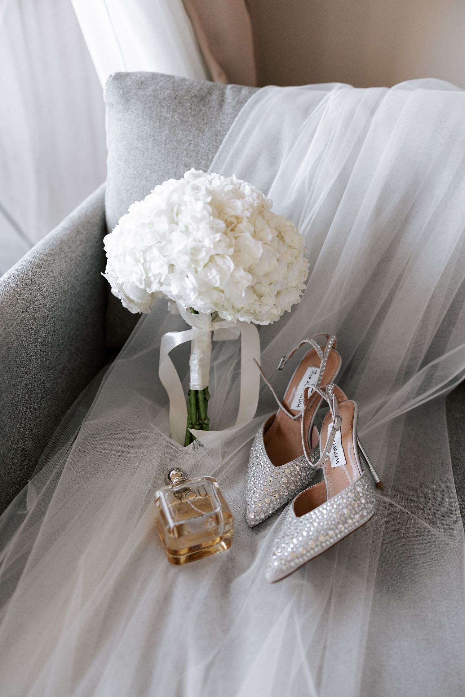 Wedding accessories including sparkly heels, white hydrangea bouquet and perfume arranged on tulle fabric.