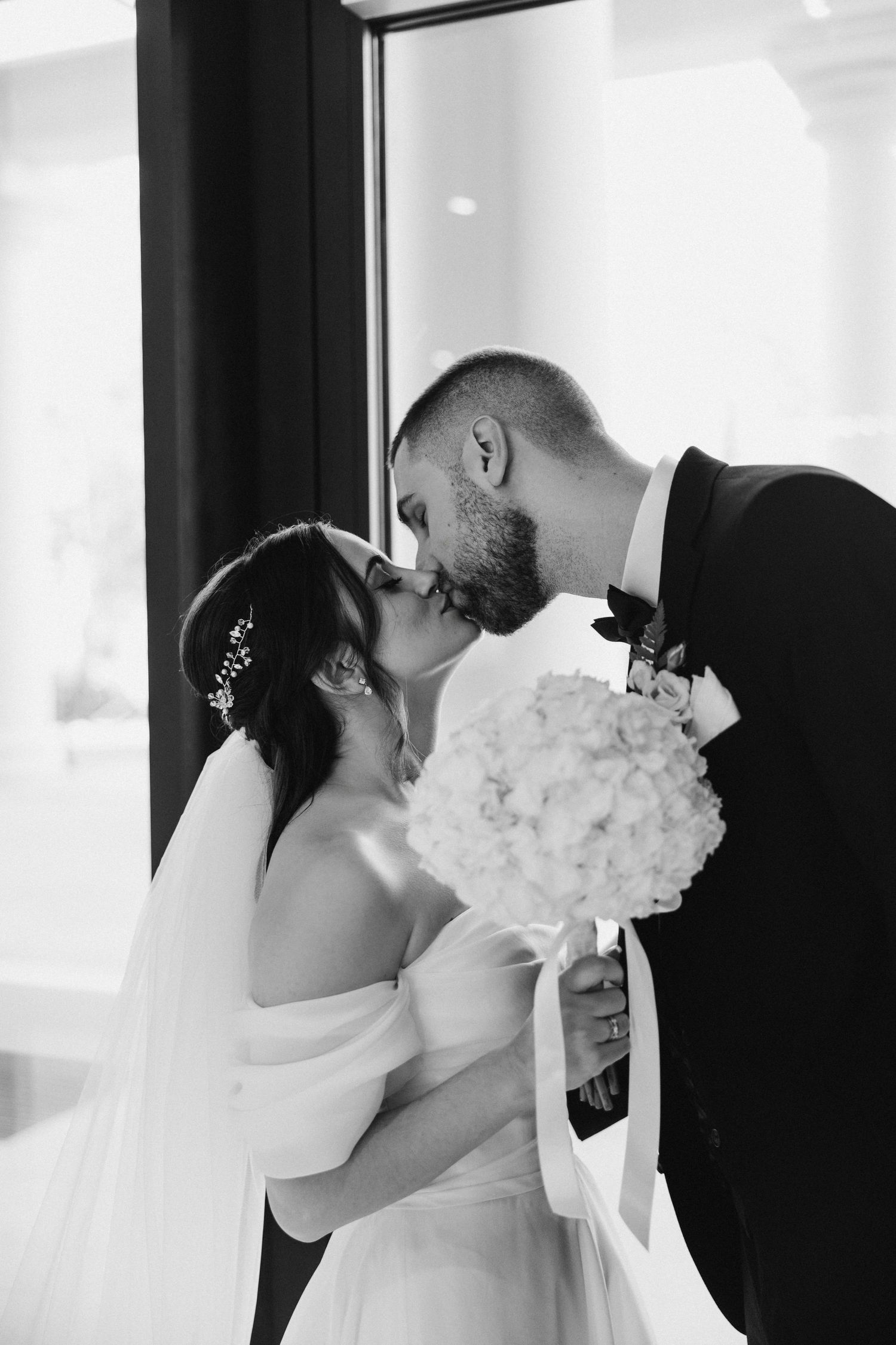 Romantic black and white wedding photo of couple kissing while holding a white floral bouquet.