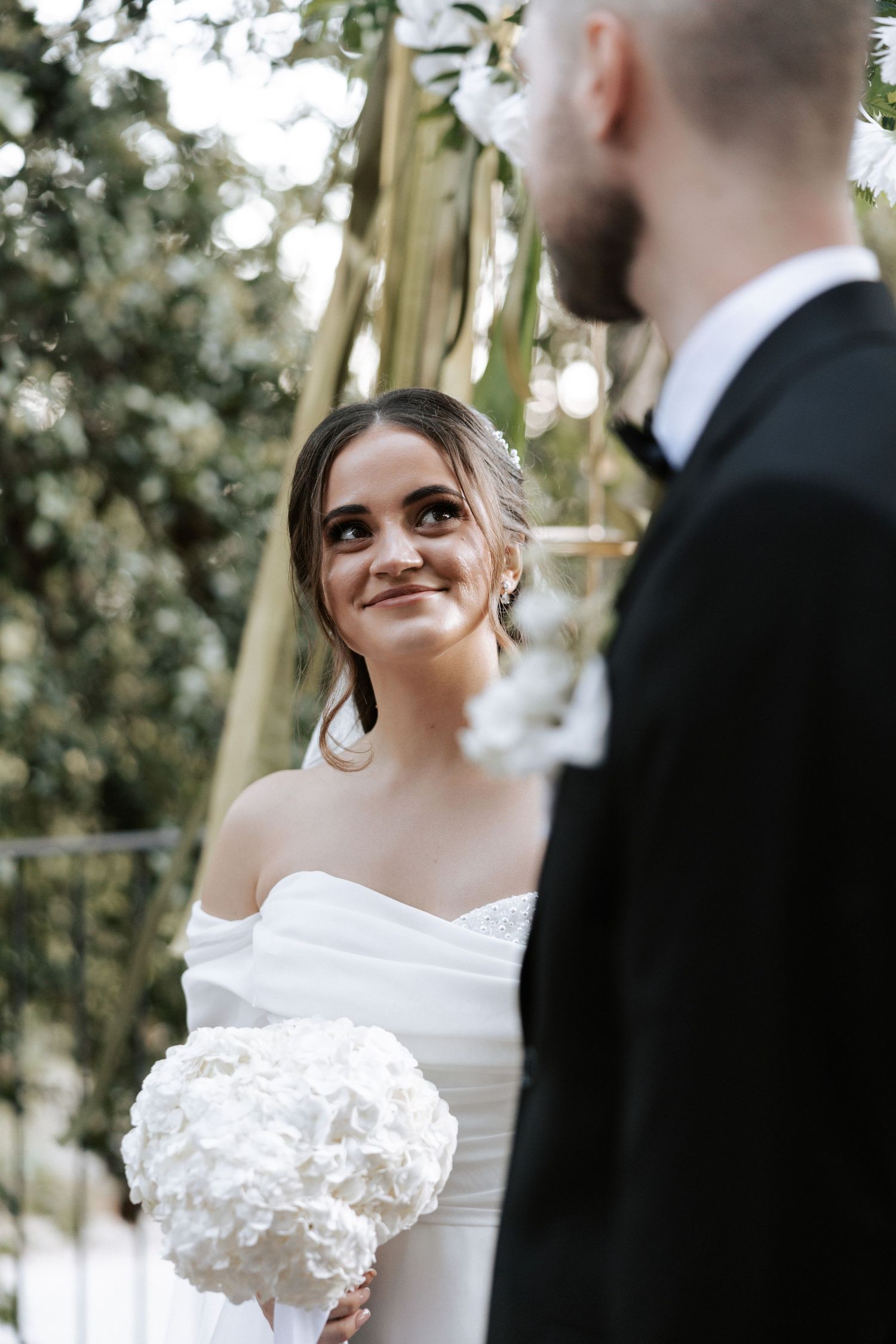 A bride in a white dress smiles lovingly while holding a white bouquet during an outdoor wedding ceremony.
