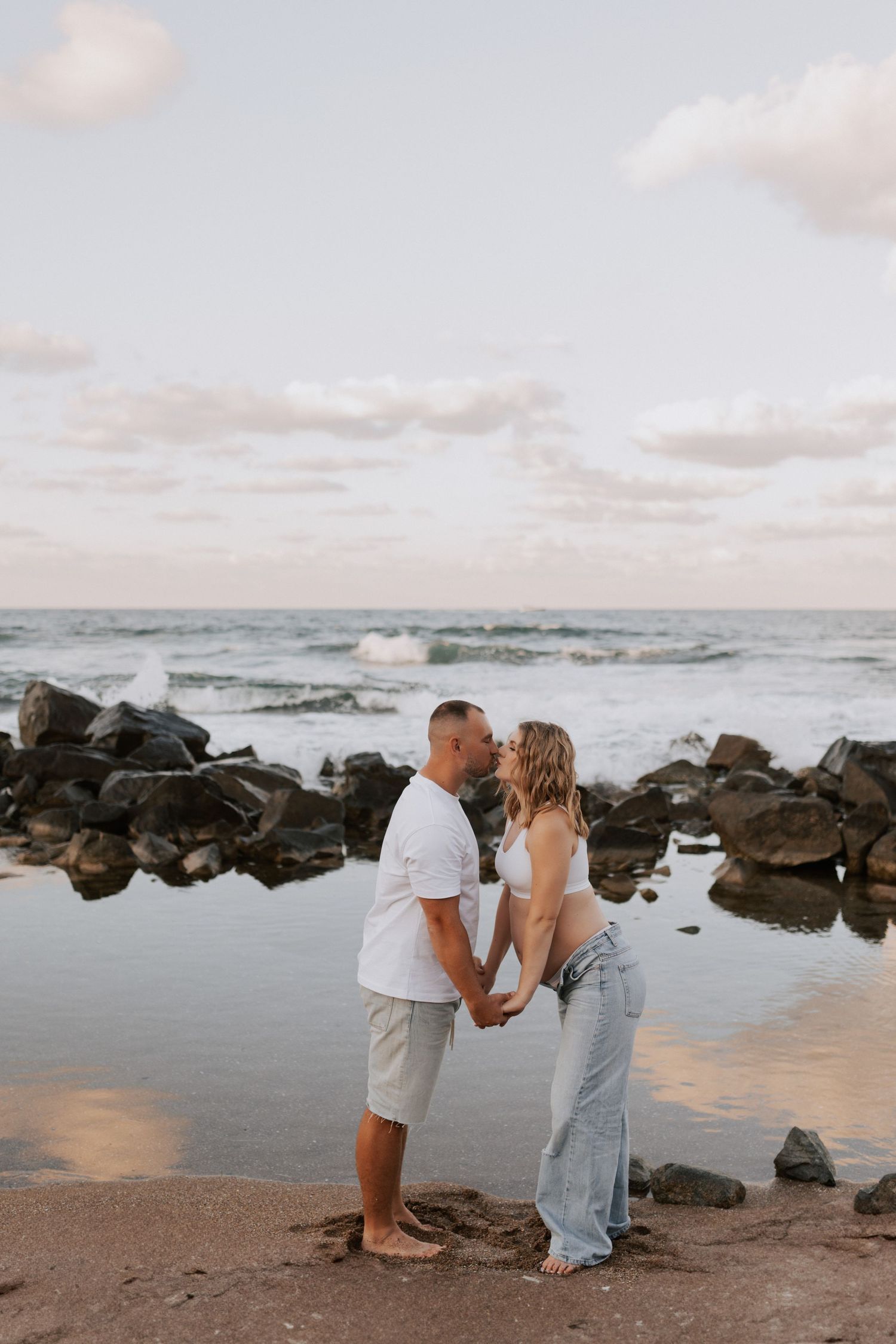 A couple holds hands while standing among rocky coastal formations at sunset.