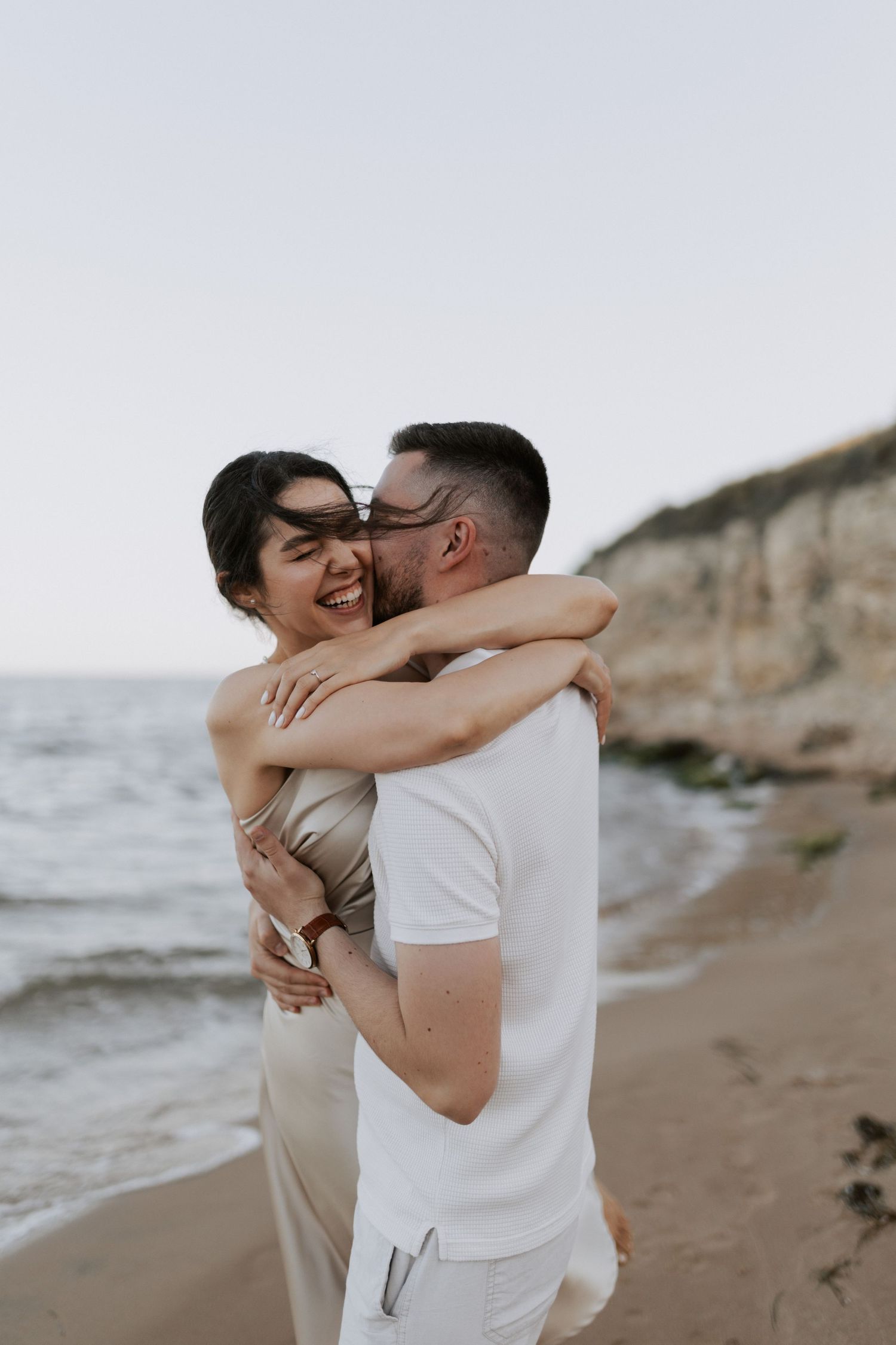 Joyful couple sharing a loving embrace on a beach with coastal cliffs behind them.