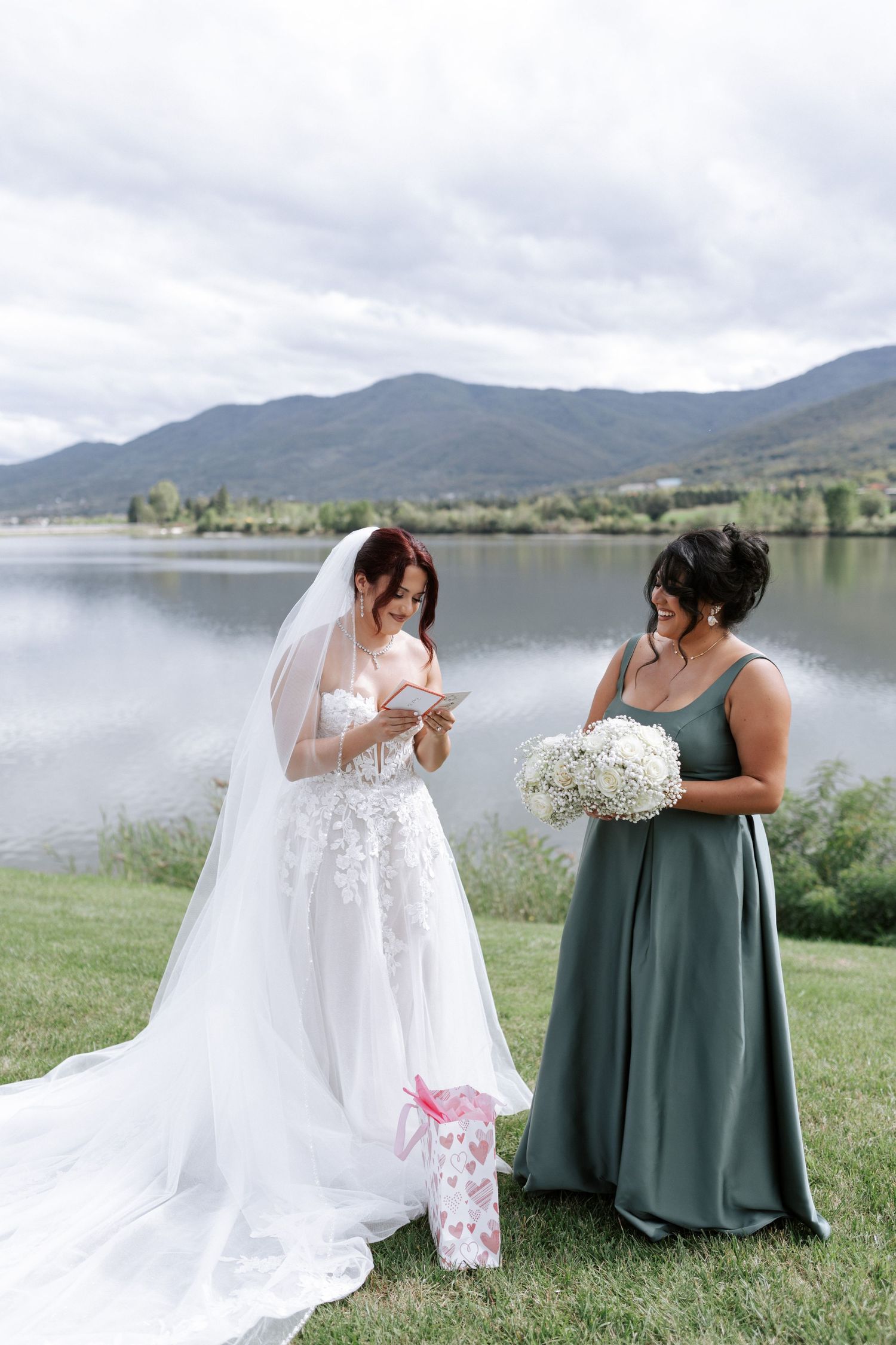Bride and bridesmaid share a moment while opening gifts against mountain lake backdrop.