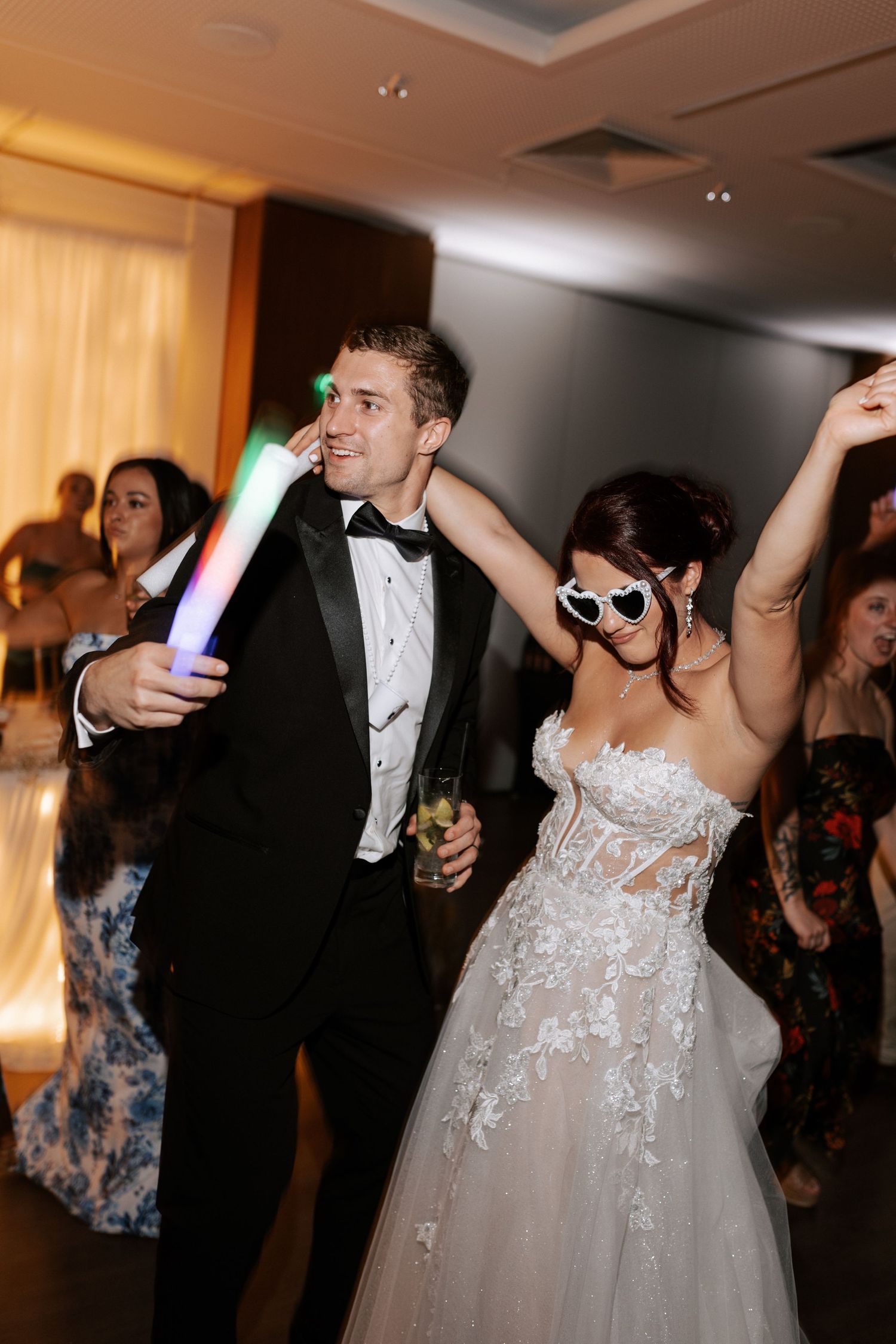 Newlyweds dancing at wedding reception with LED light sticks and sunglasses in elegant formal attire.