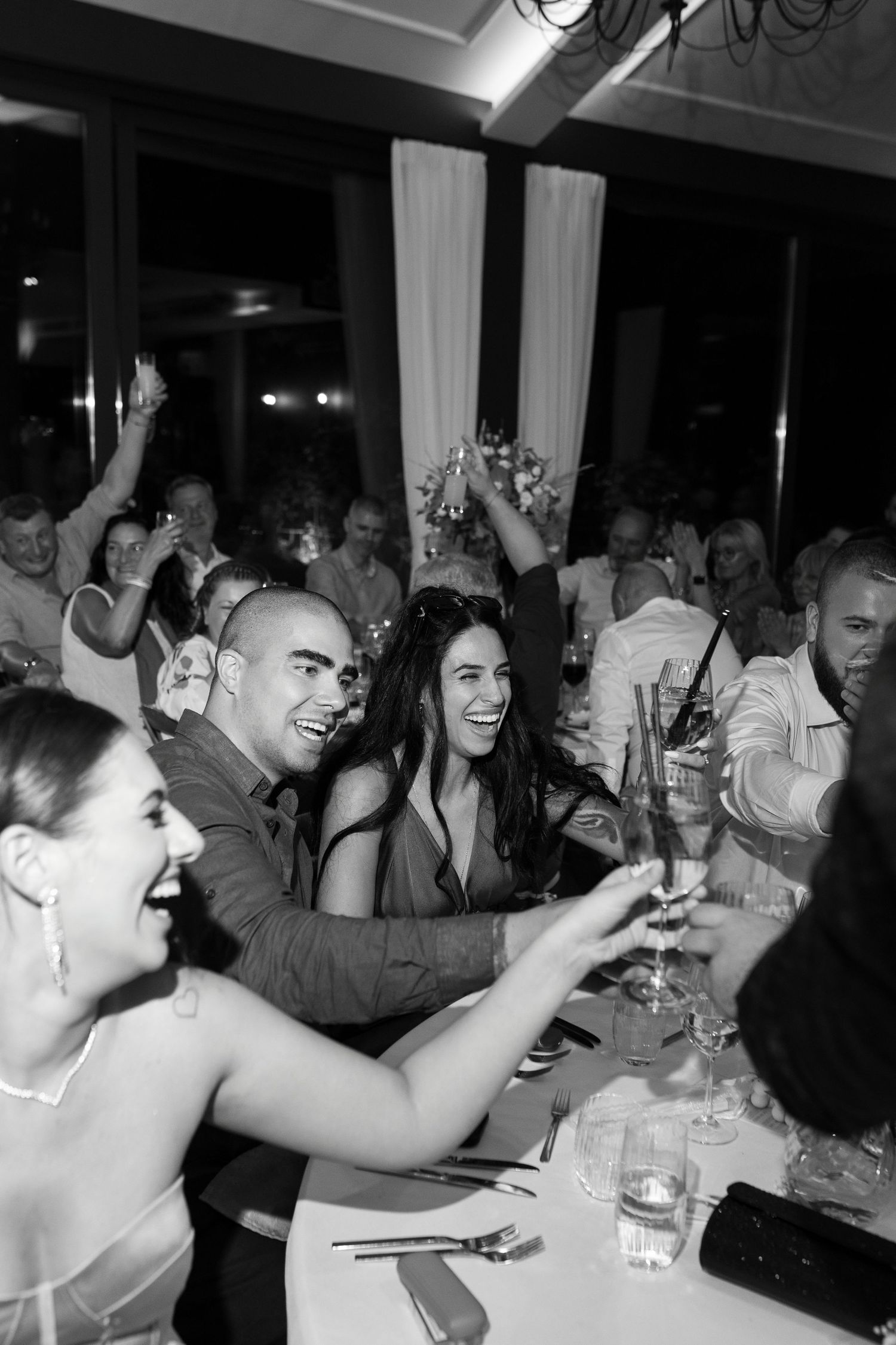Party guests raise their glasses in celebration at a lively indoor event with white drapes and dim lighting.