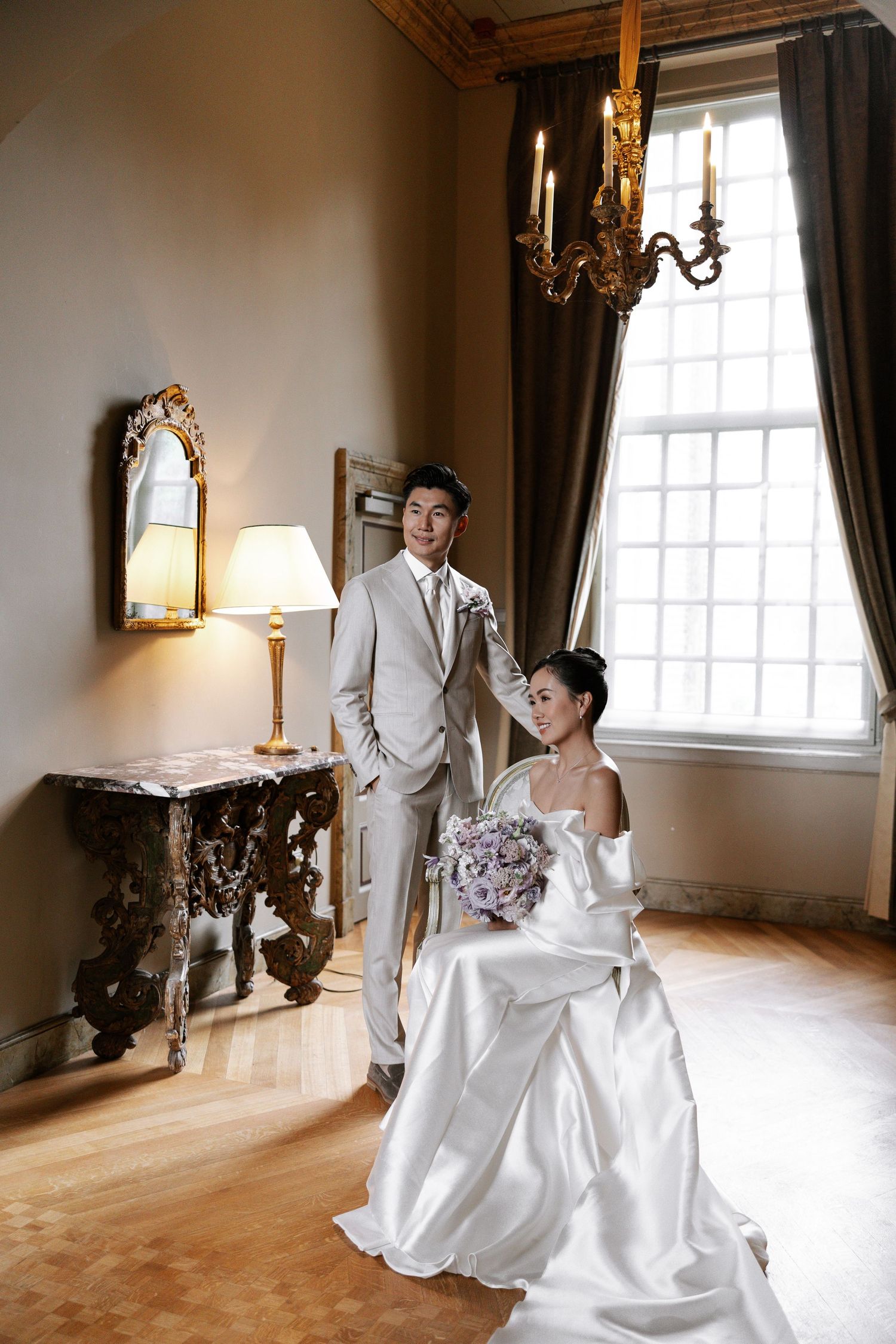 Elegant wedding couple poses in a vintage room with hardwood floors, ornate mirrors, and natural window light.